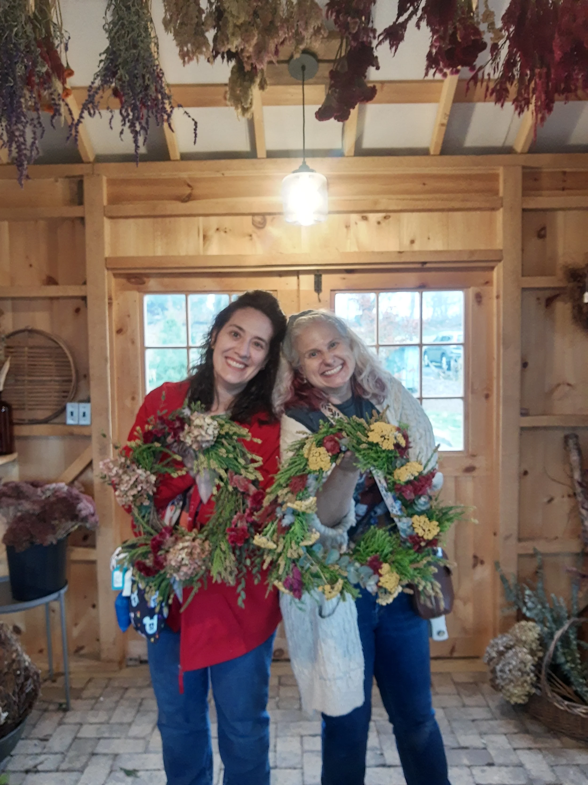 Two women standing inside a wooden shop, smiling and holding floral wreaths made of autumn flowers and greenery.