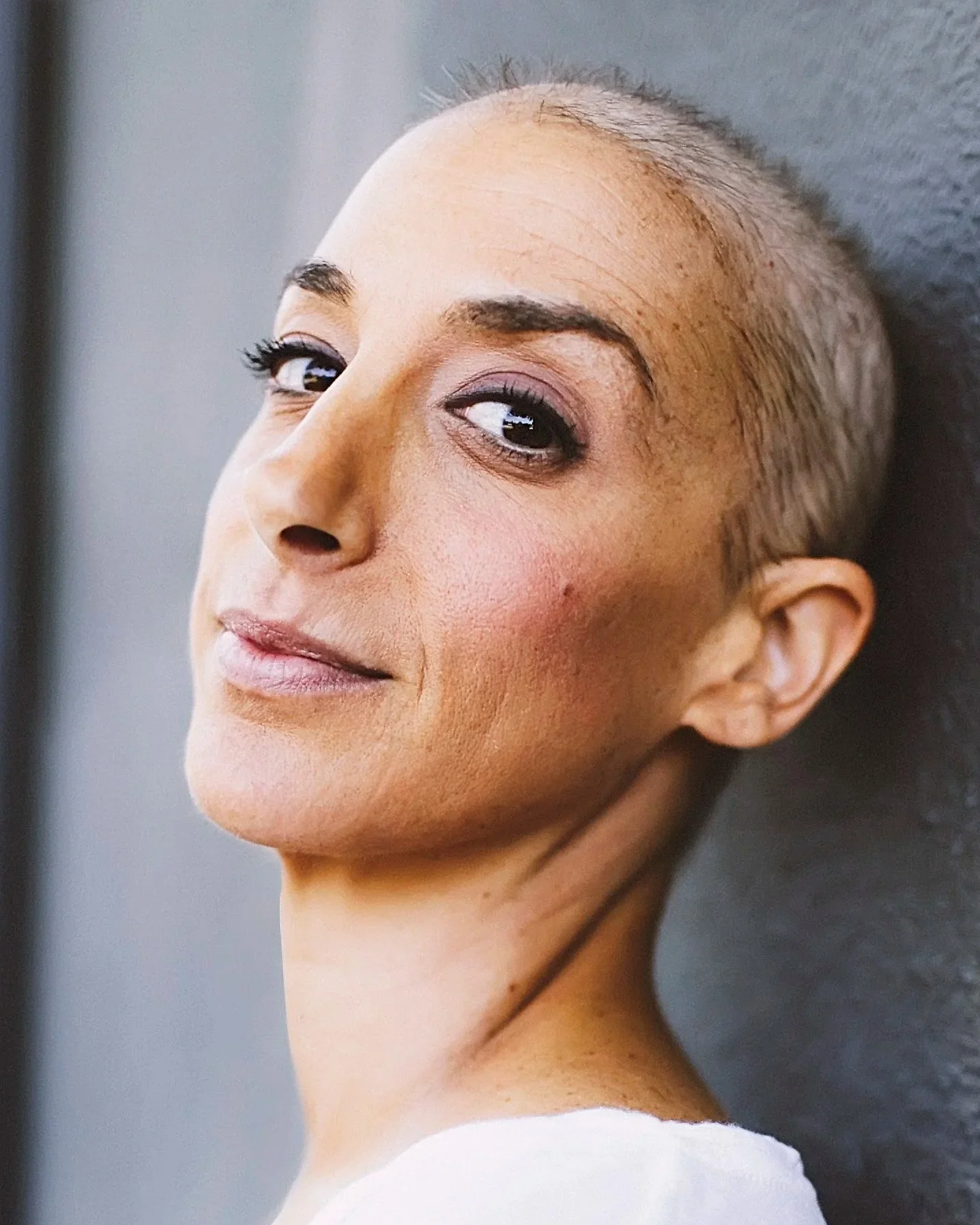 Close-up of a woman with a shaved head, wearing makeup, lying on a textured dark gray surface, and looking over her shoulder.