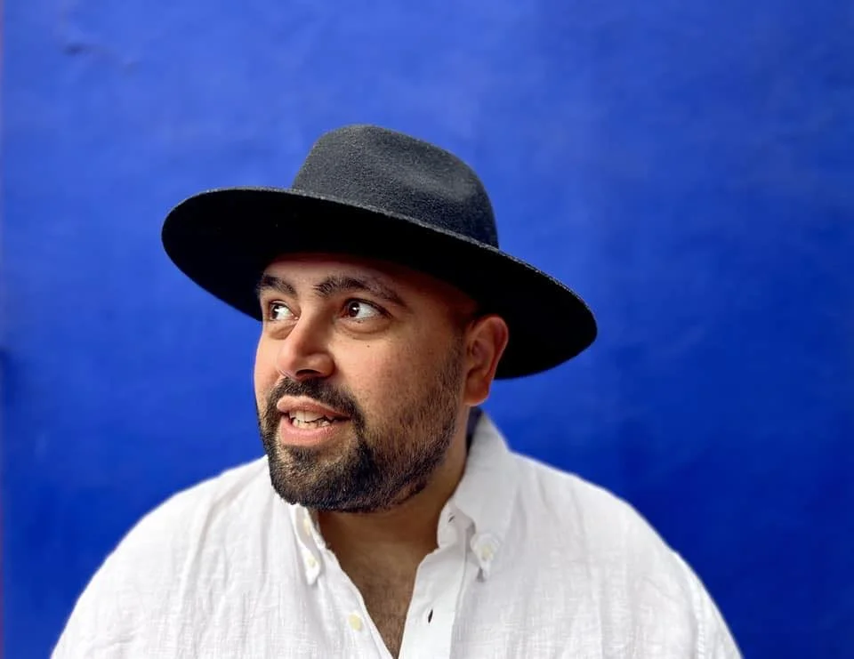 Man with a beard wearing a white shirt and a black wide-brimmed hat against a blue background.