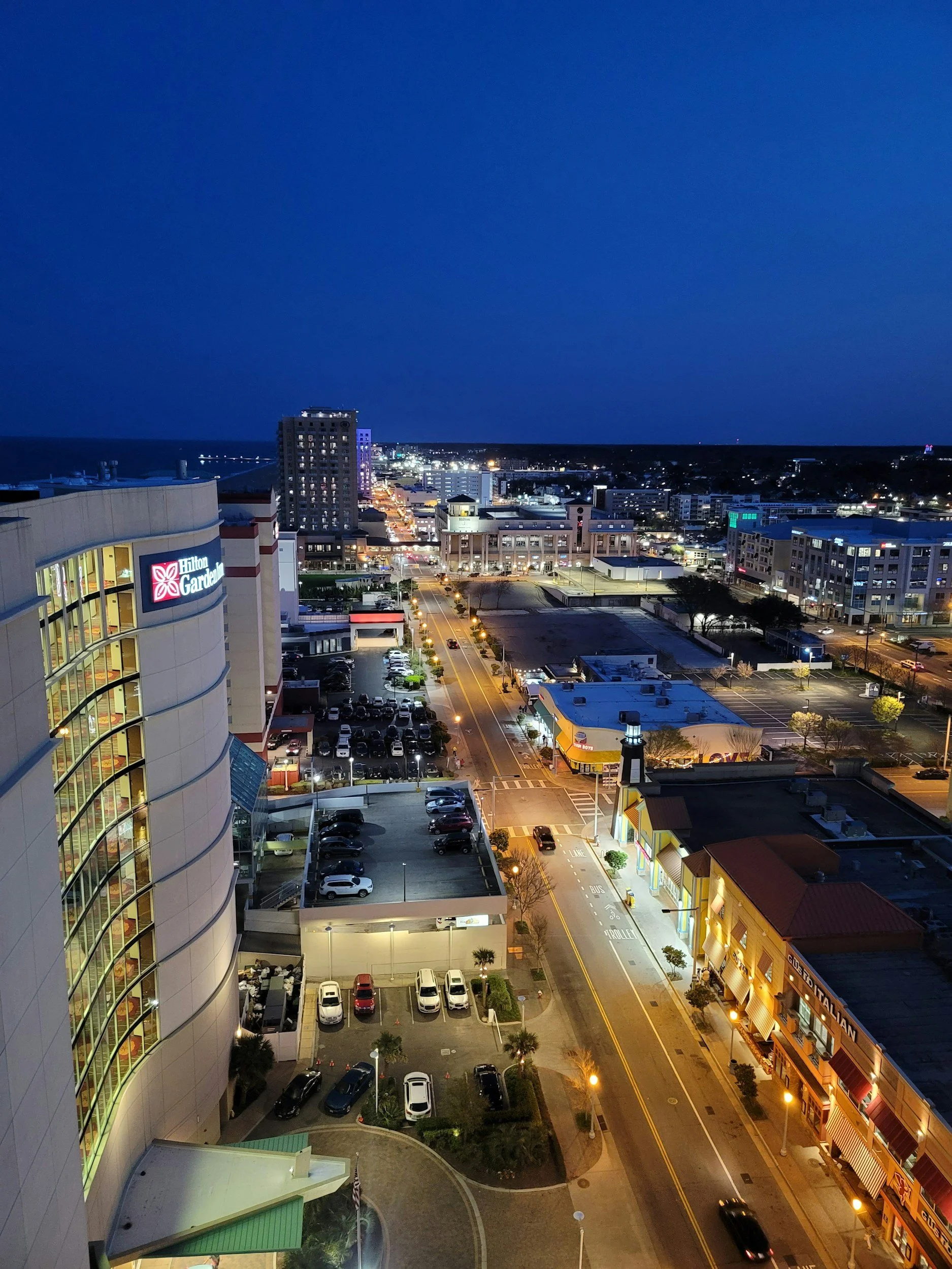 Aerial view of a cityscape at night with illuminated streets and buildings, including a Hilton Garden Inn sign. There are parked cars along the streets and in parking lots. The scene captures a vibrant urban environment under a dark evening sky.