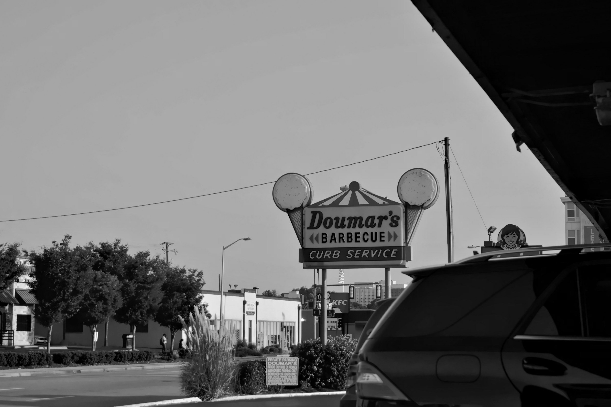 Black and white photo of Doumar's Barbecue sign with curb service, trees, parked cars, and adjacent fast-food signs.