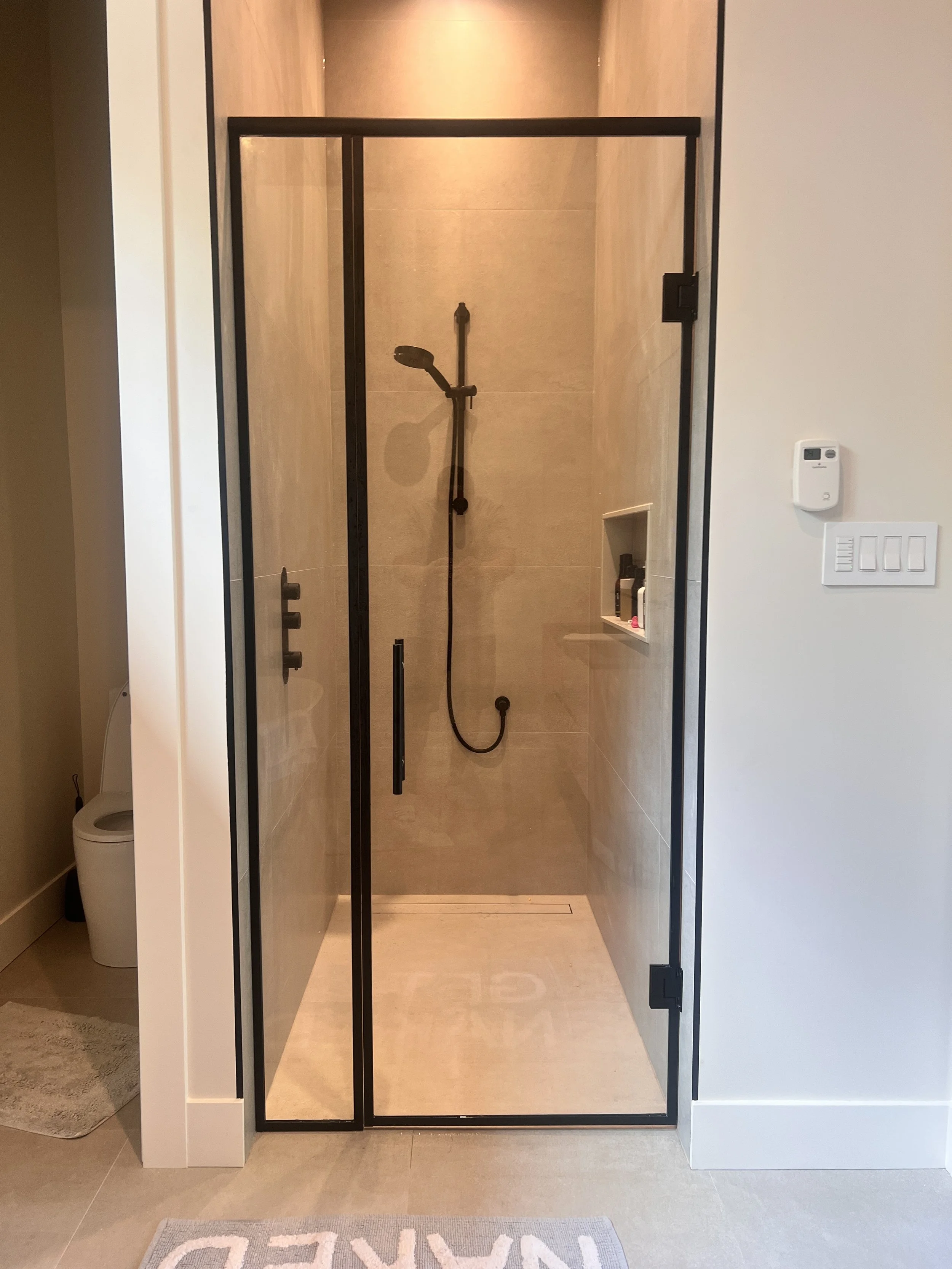 A modern shower enclosure with black metal framing, inside beige tiled walls, a handheld showerhead, built-in shelf containing toiletries, and a floor drain.