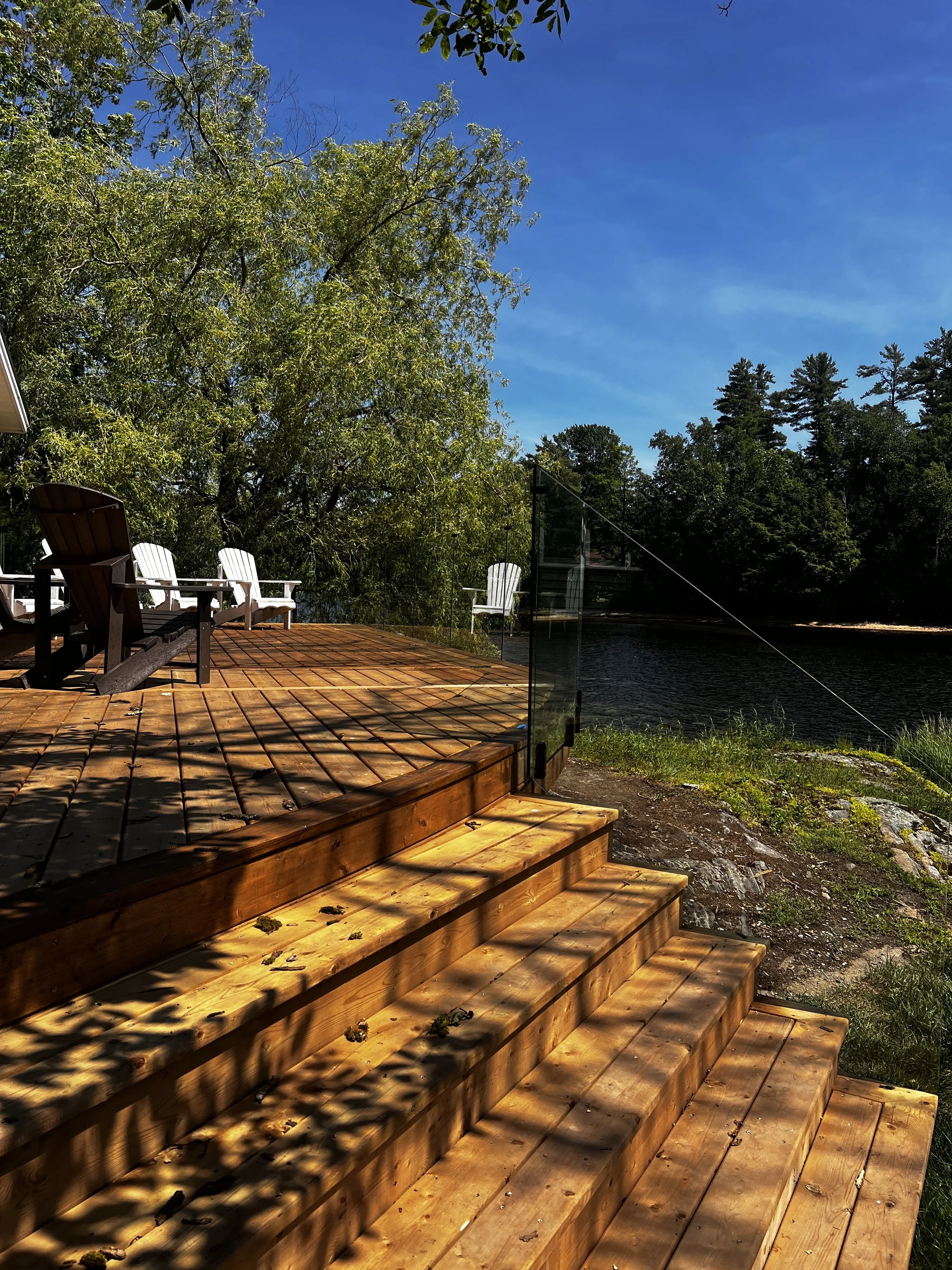 A wooden deck with stairs leading down to a grassy area by the water, with trees and a blue sky in the background. There are white chairs on the deck, and a glass barrier on one side.