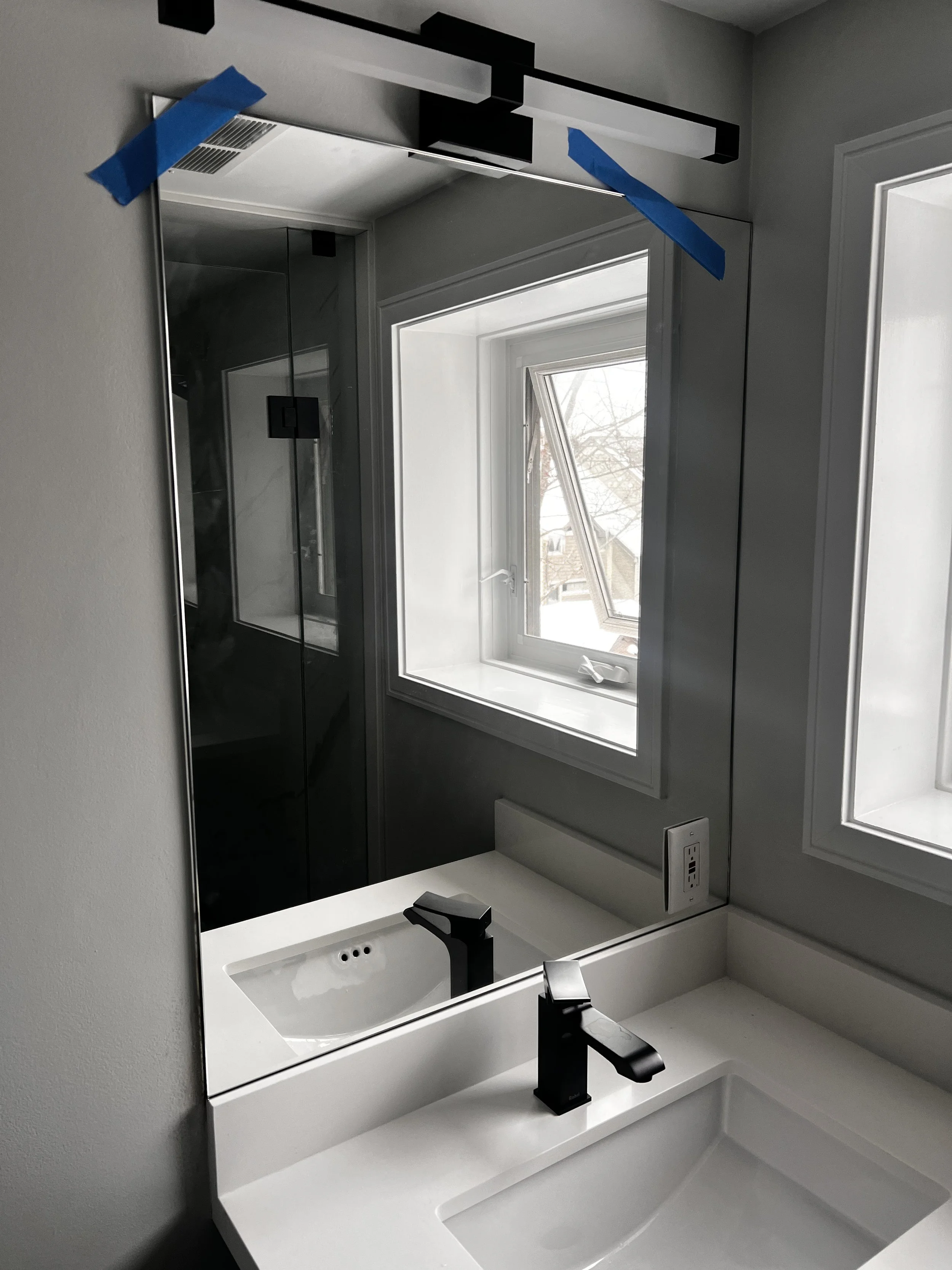 Bathroom with a modern white sink, black faucet, large mirror above, window to the right, and an electrical outlet on the wall, with reflections visible in the mirror.