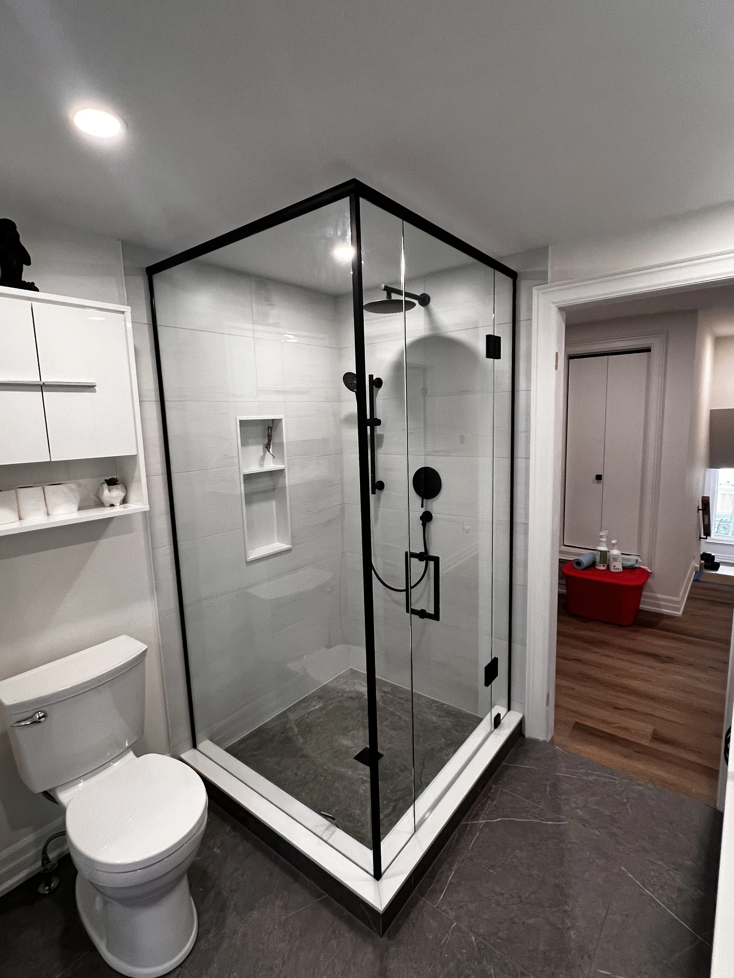 A modern bathroom featuring a glass walk-in shower with black framing, white tiled walls, and a built-in niche. Adjacent to the shower is a toilet, and to the right, an open doorway reveals a room with wooden flooring and a red storage bin.