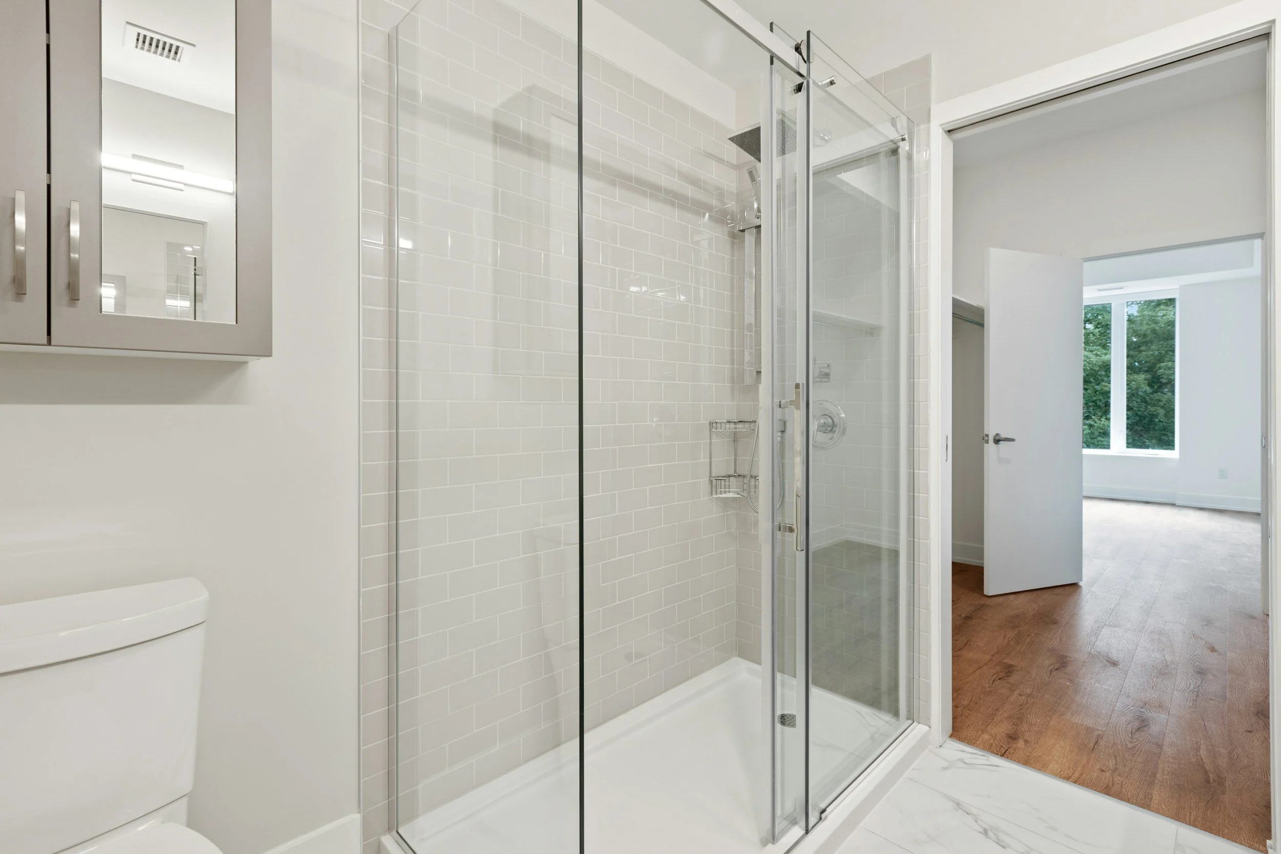 Modern bathroom with a glass shower enclosure, white tiled walls, a white toilet, a gray cabinet with mirrored doors, and a view into a bright room with large windows and wooden flooring.