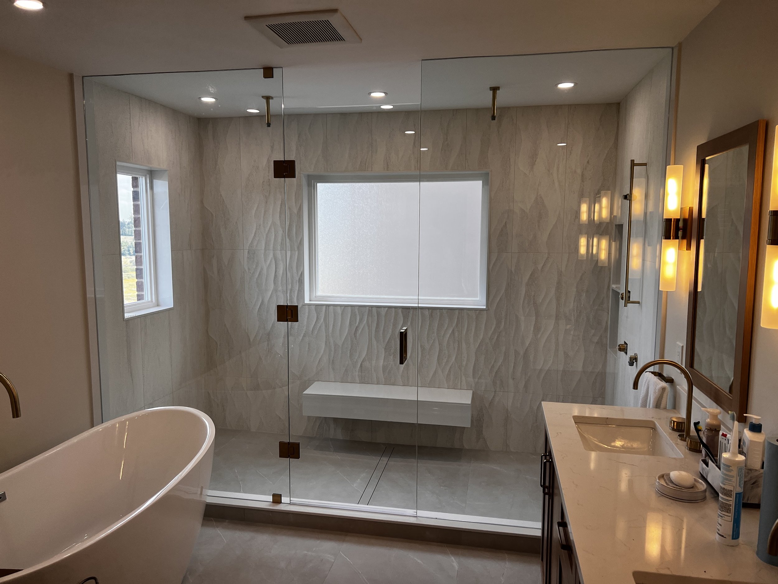 Modern bathroom with glass shower enclosure, bathtub, window, and vanity with sink, mirror, and lighting fixtures.