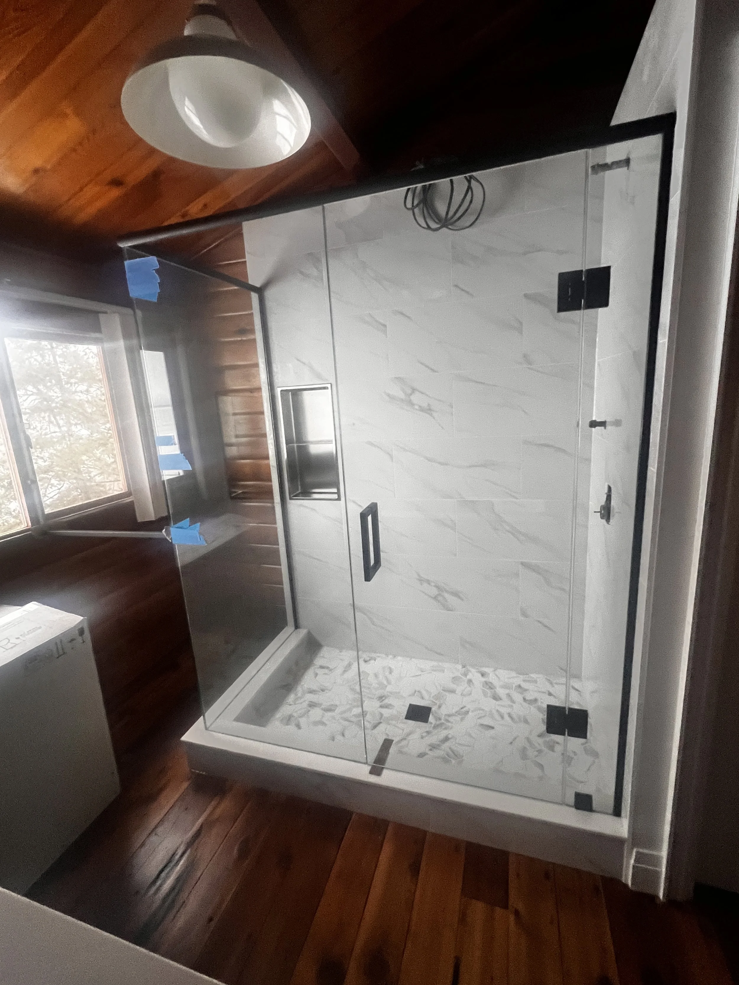 A glass shower enclosure with marble wall tiles and a pebble floor, located in a room with wooden paneling and a window.