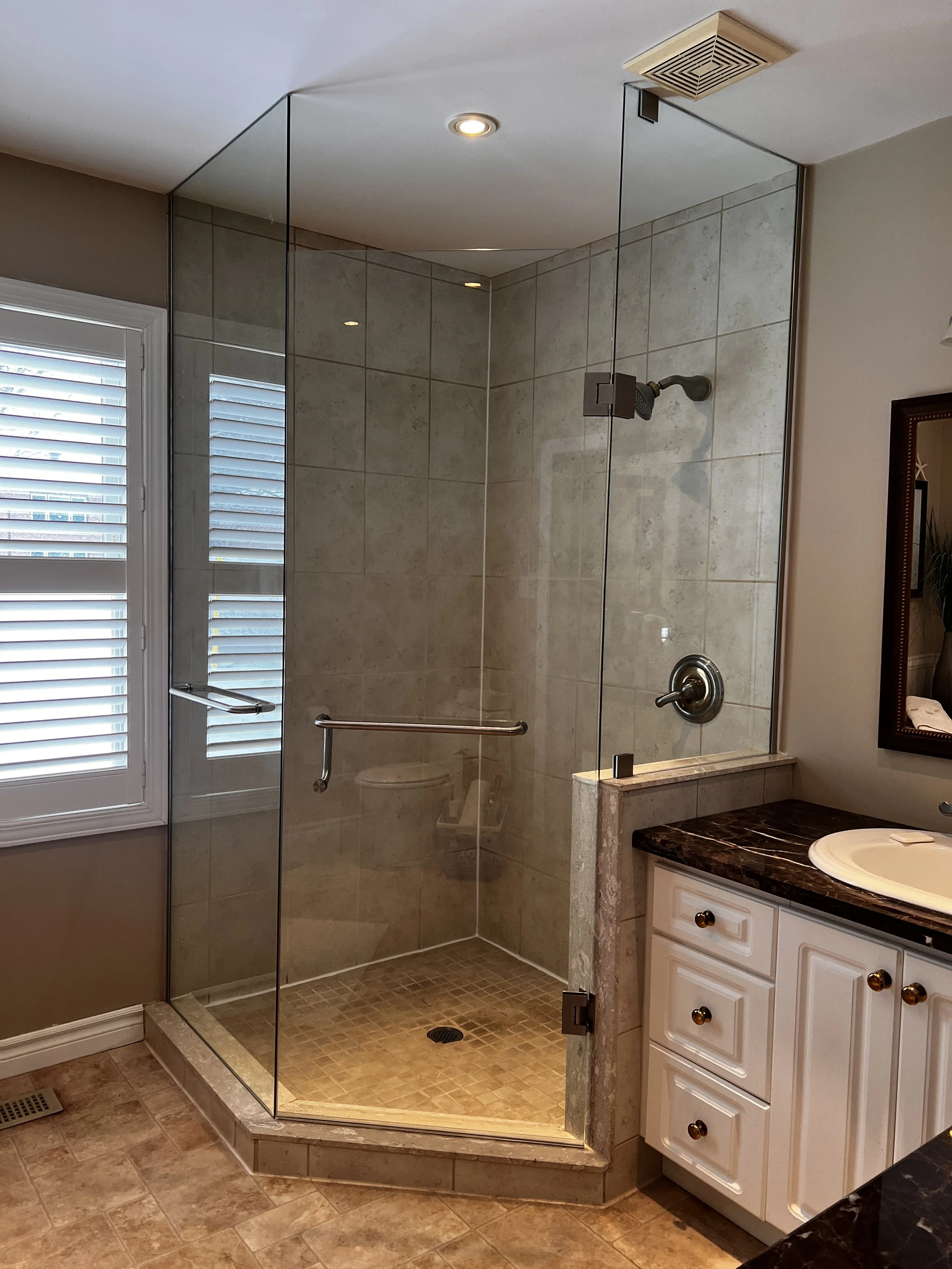 A bathroom with a walk-in glass shower, beige tiled walls and floor, a window with blinds, a white bathroom vanity with a dark marble countertop, and a mirror on the wall.