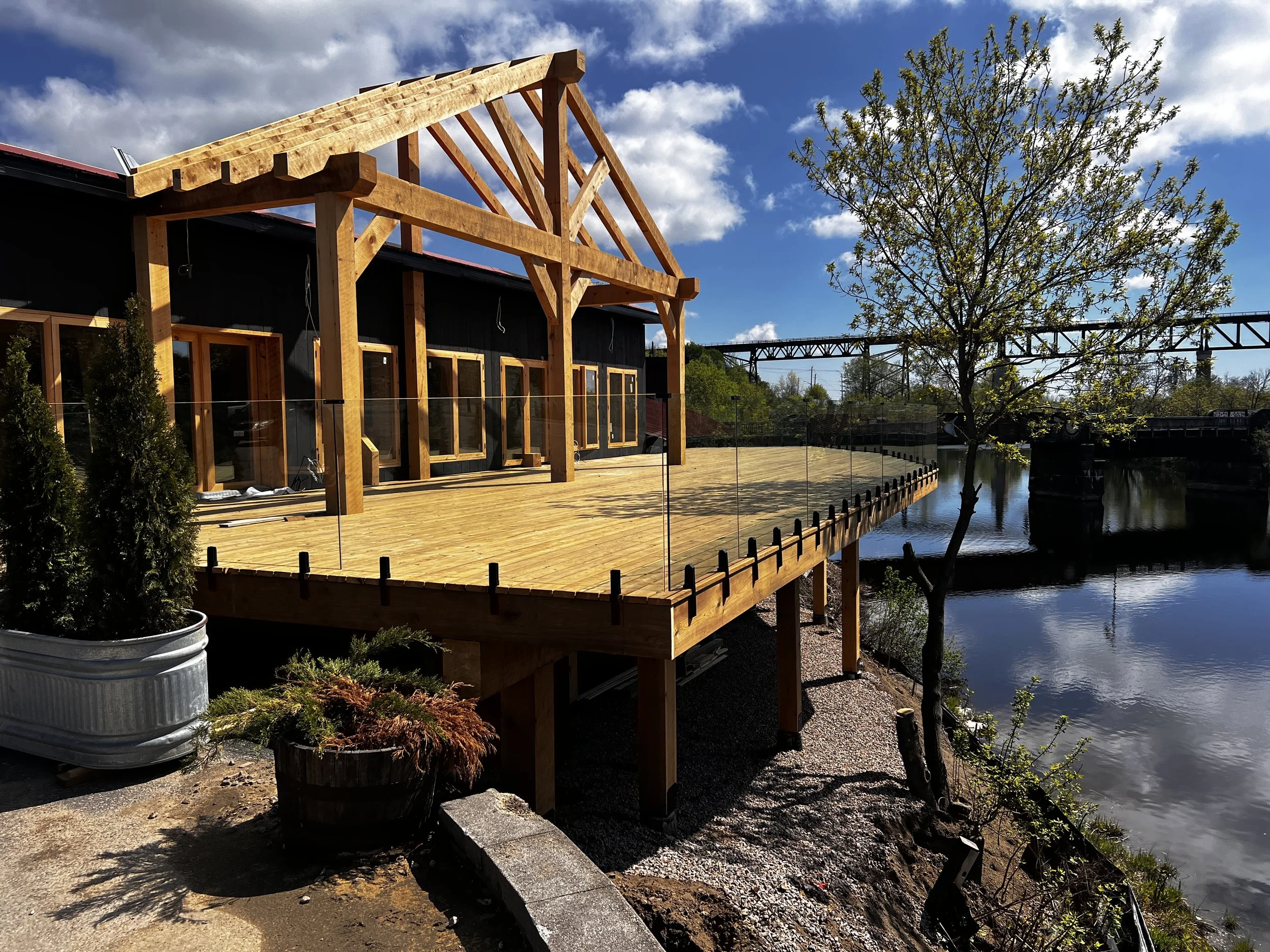 Construction of a wooden porch or deck next to a river, with a tree and a metal railing on the waterside, under a partly cloudy sky.