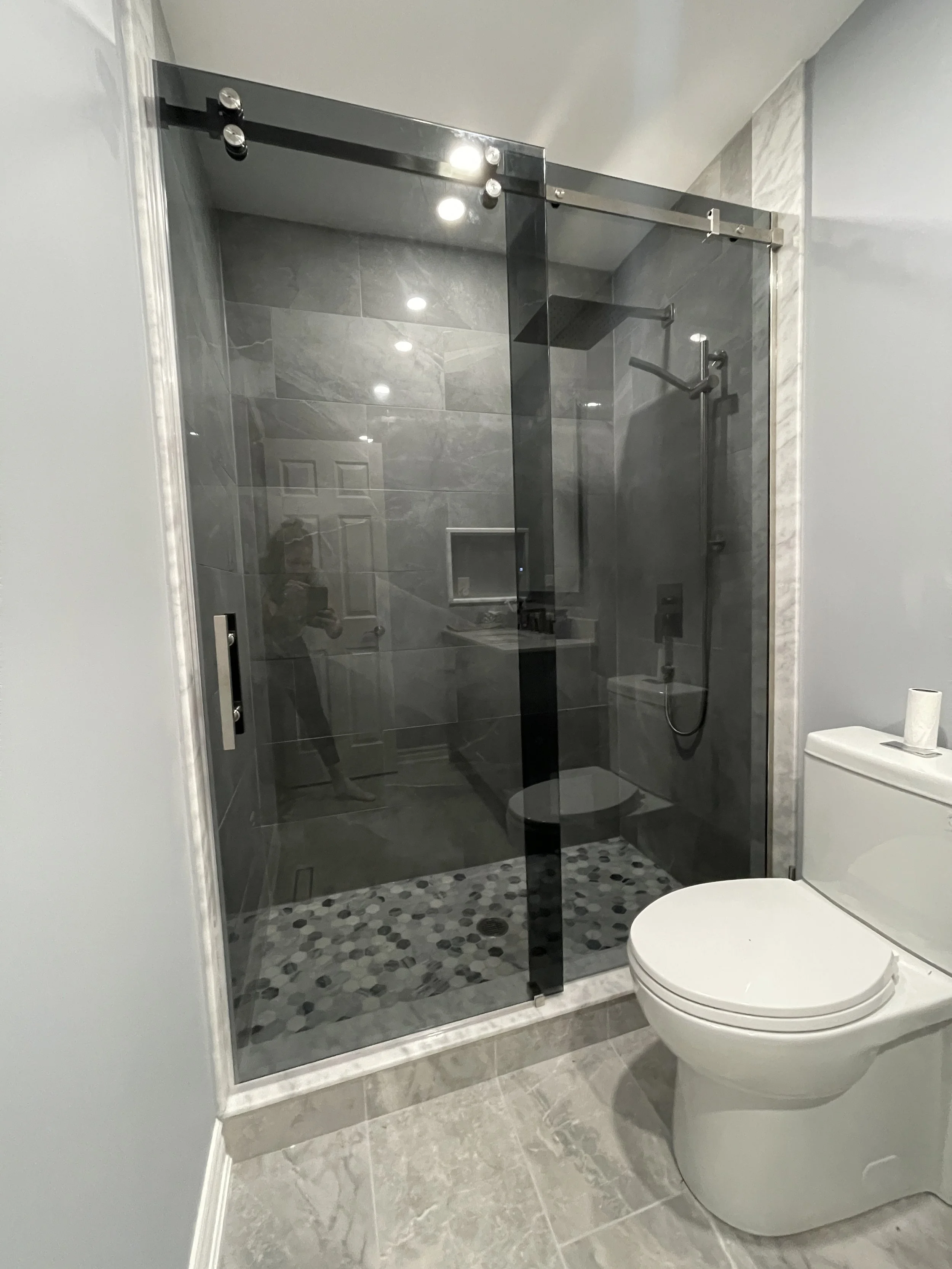 A bathroom with a glass shower enclosure and a white toilet. The shower has gray tiles and a pebble-patterned shower floor. The bathroom floor is tiled with gray tiles.