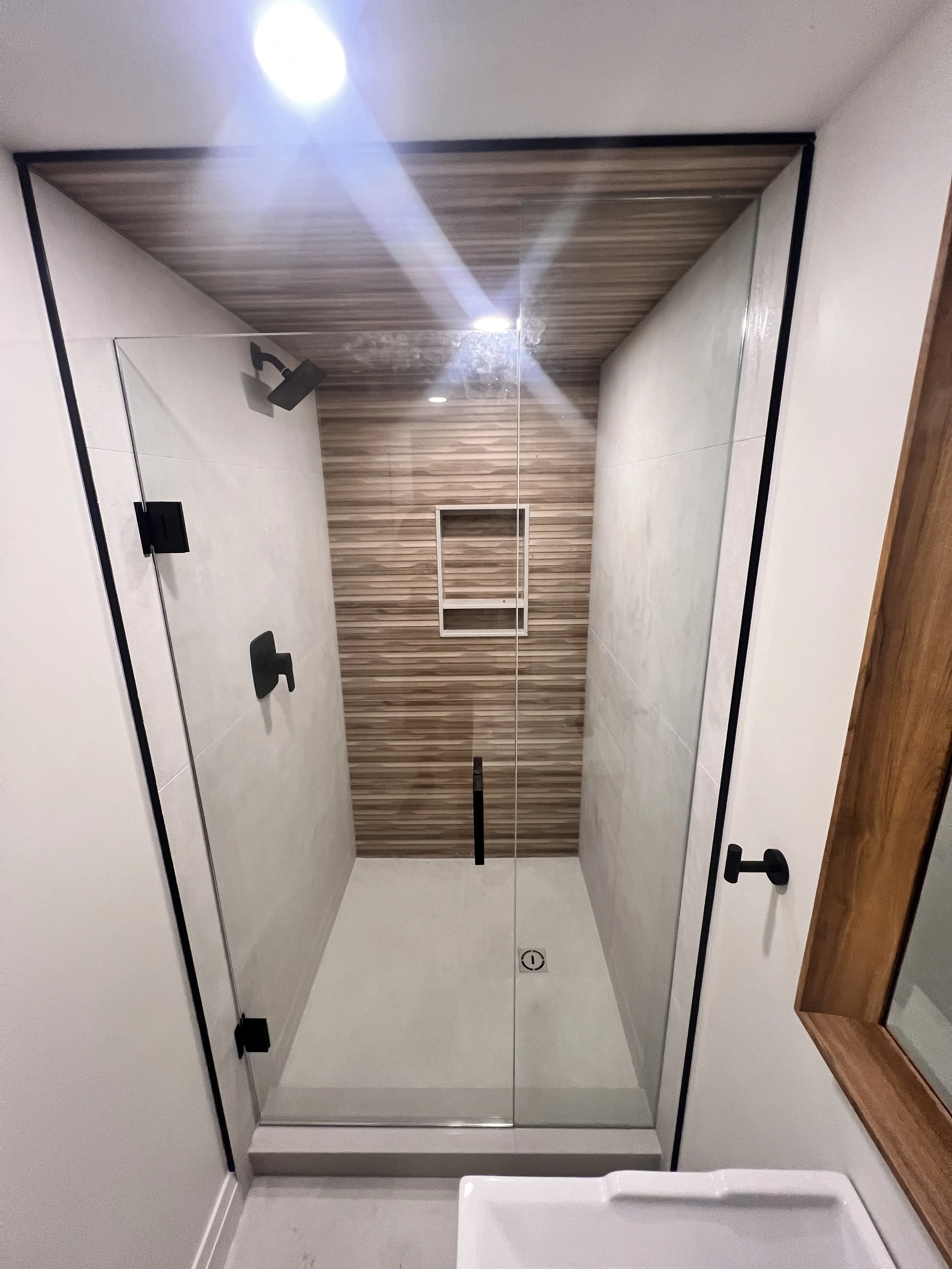 Modern walk-in shower with glass door, black fixtures, wooden ceiling and wall accent, and built-in shelf in the back wall.