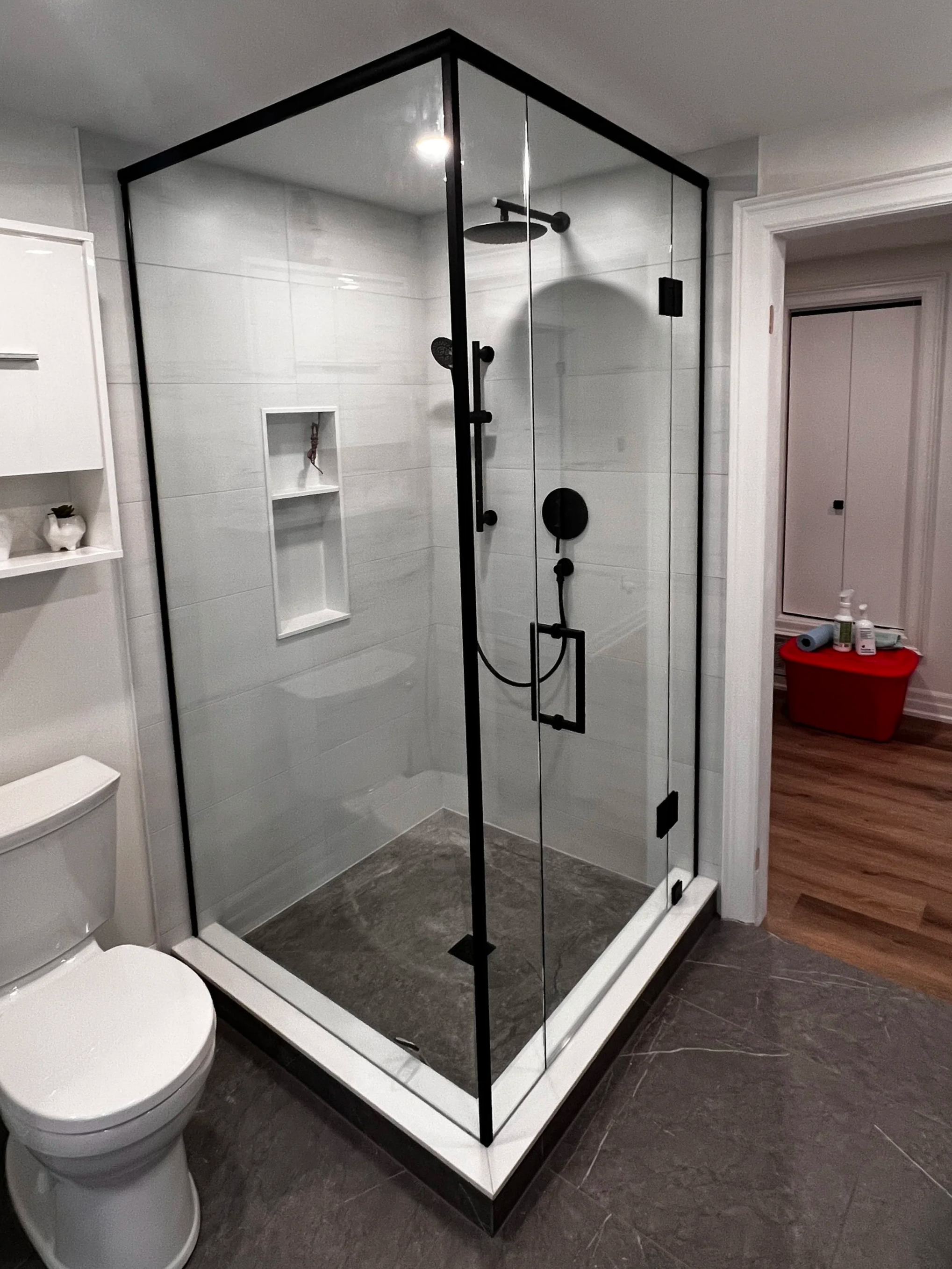 A modern glass shower enclosure with black hardware in a bathroom, with a toilet and storage shelves nearby.
