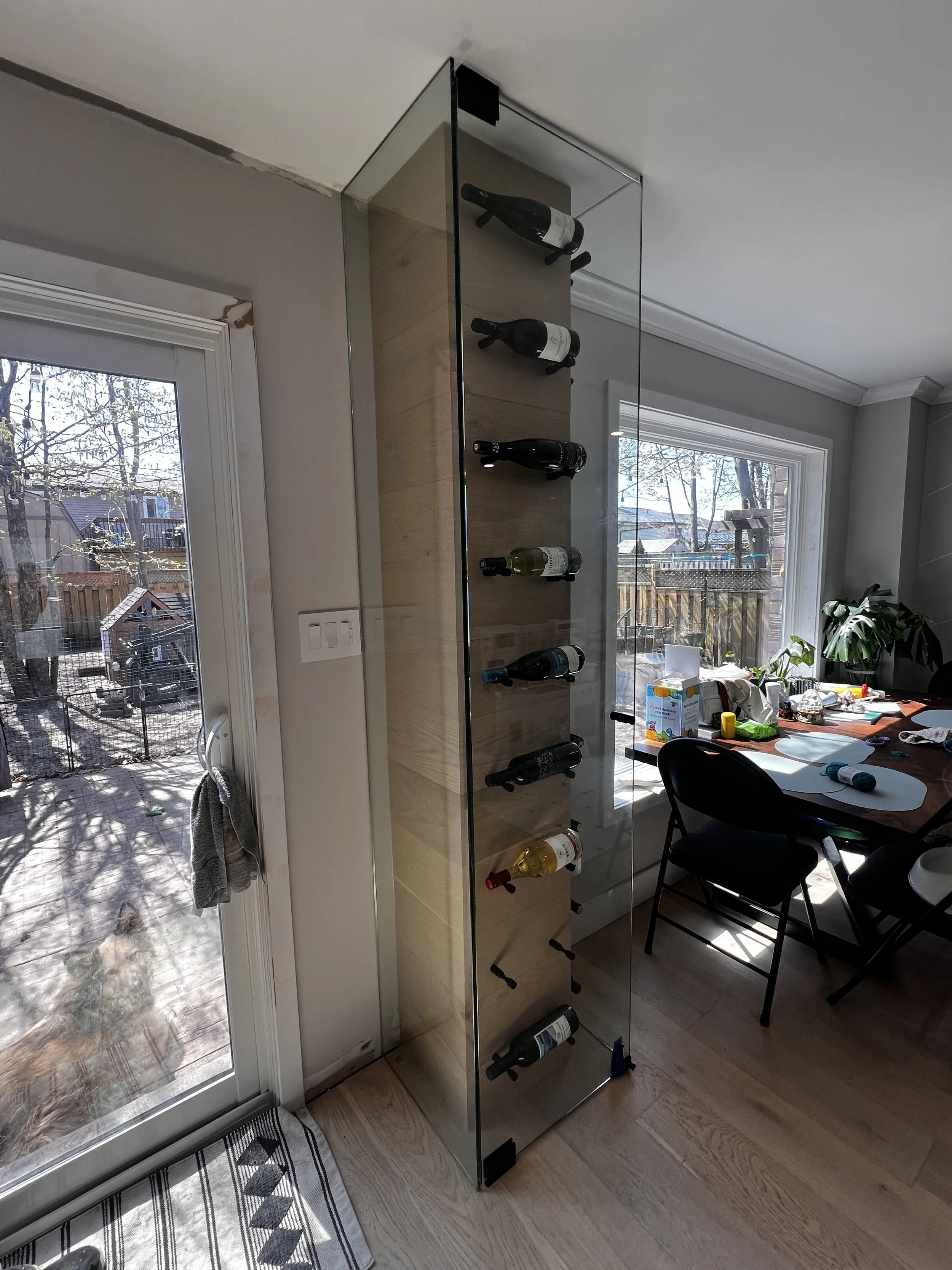 A glass wine rack with a wooden back panel holding nine bottles of wine, situated inside a room near a sliding glass door that opens to a backyard with trees and a dog house.
