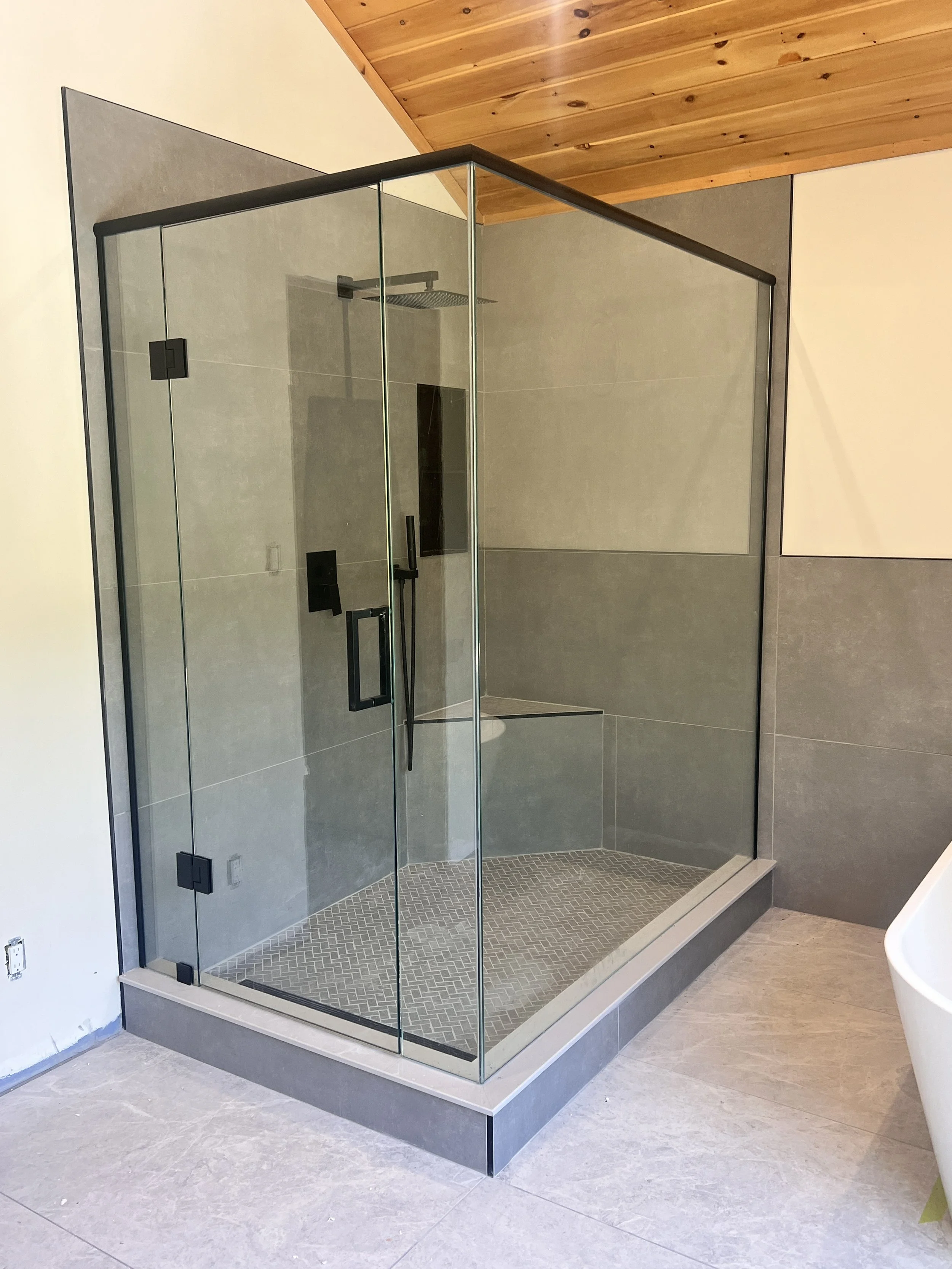 Glass shower enclosure with black hardware, installed in a modern bathroom with a wood-paneled ceiling and neutral gray tiled walls and floor.