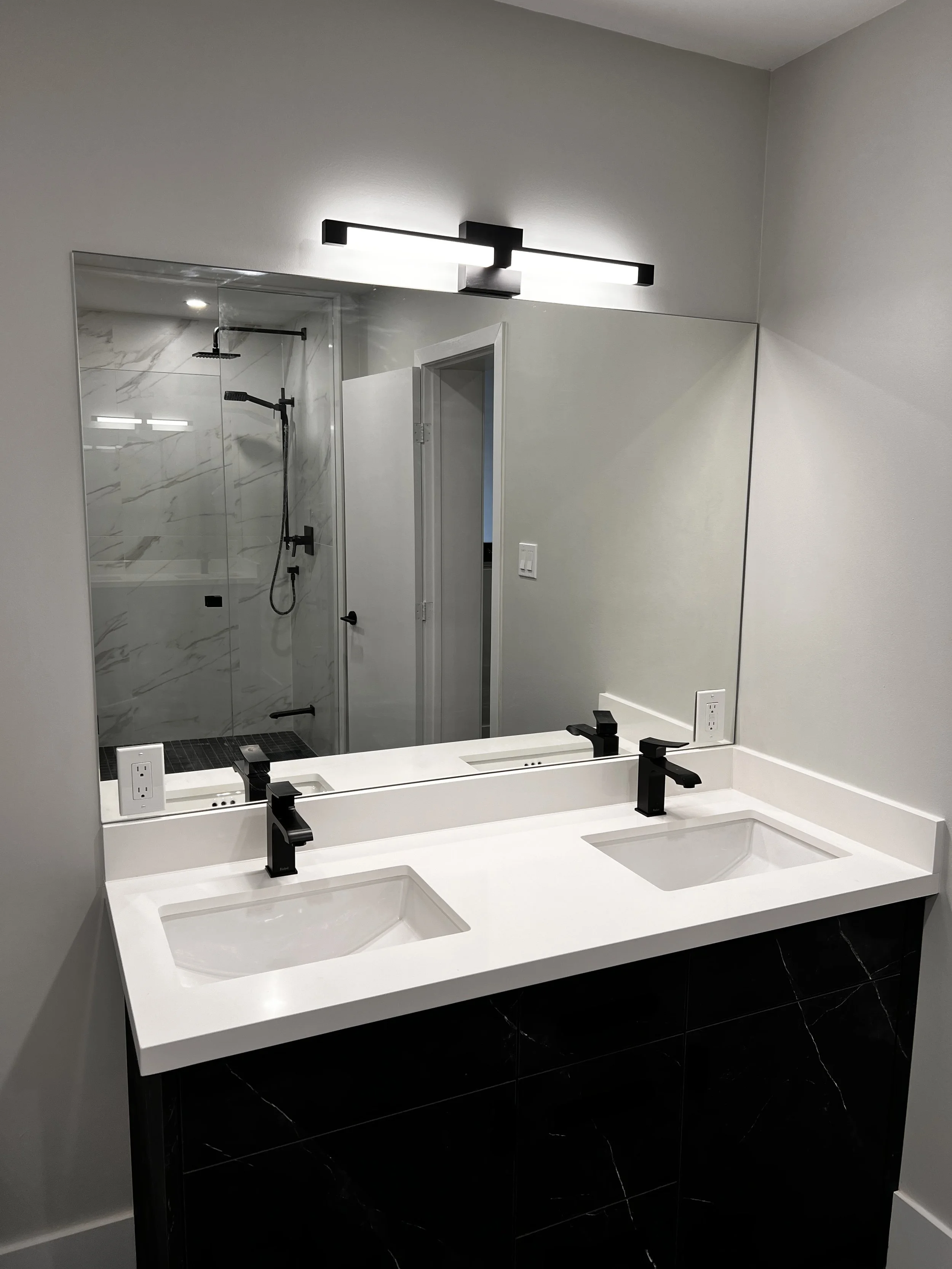 Modern bathroom with double white sink vanity, black faucets, large mirror, and walk-in shower with marble tiles and black fixtures.