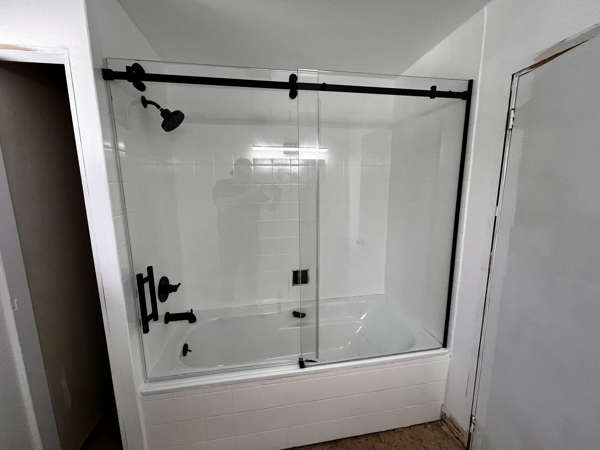 A partially assembled frameless glass shower enclosure with black hardware, installed in a white-tiled bathroom, next to a white door, with a person taking the photo reflected in the glass.