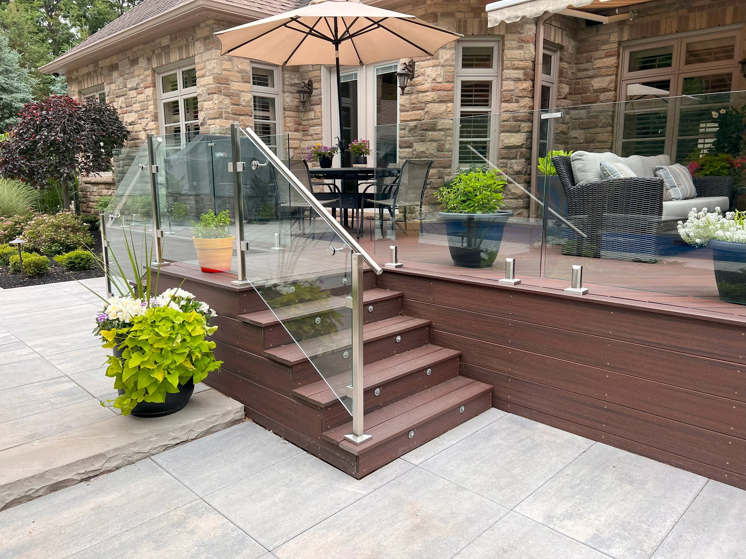 A house's backyard patio with wooden stairs, a glass railing, potted plants, outdoor furniture including a table with an umbrella and a sofa with pillows, and landscaped garden with plants and shrubs.