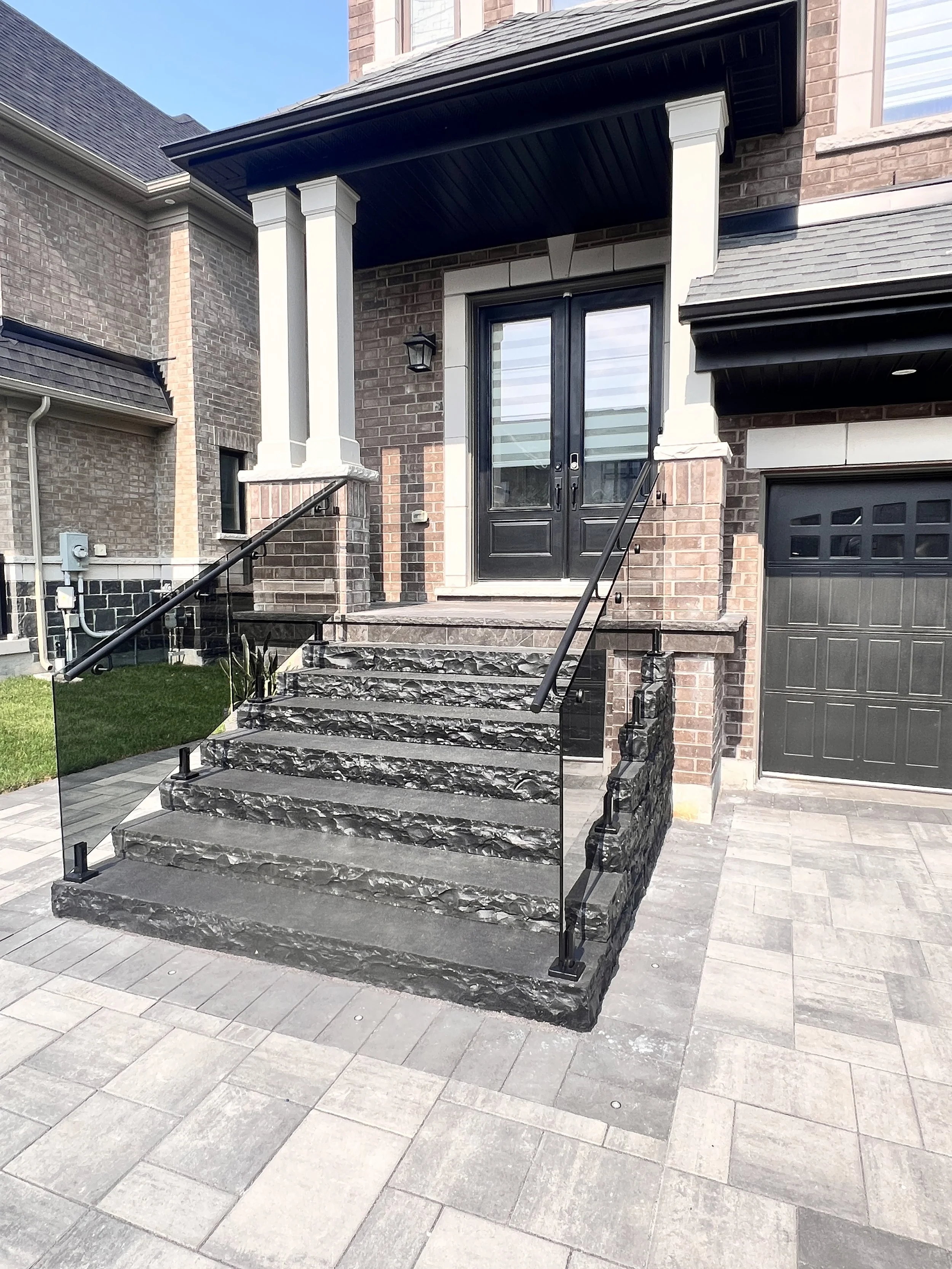 Front porch with black double doors, brick exterior, and stone steps with glass railing leading to the entrance of a house.