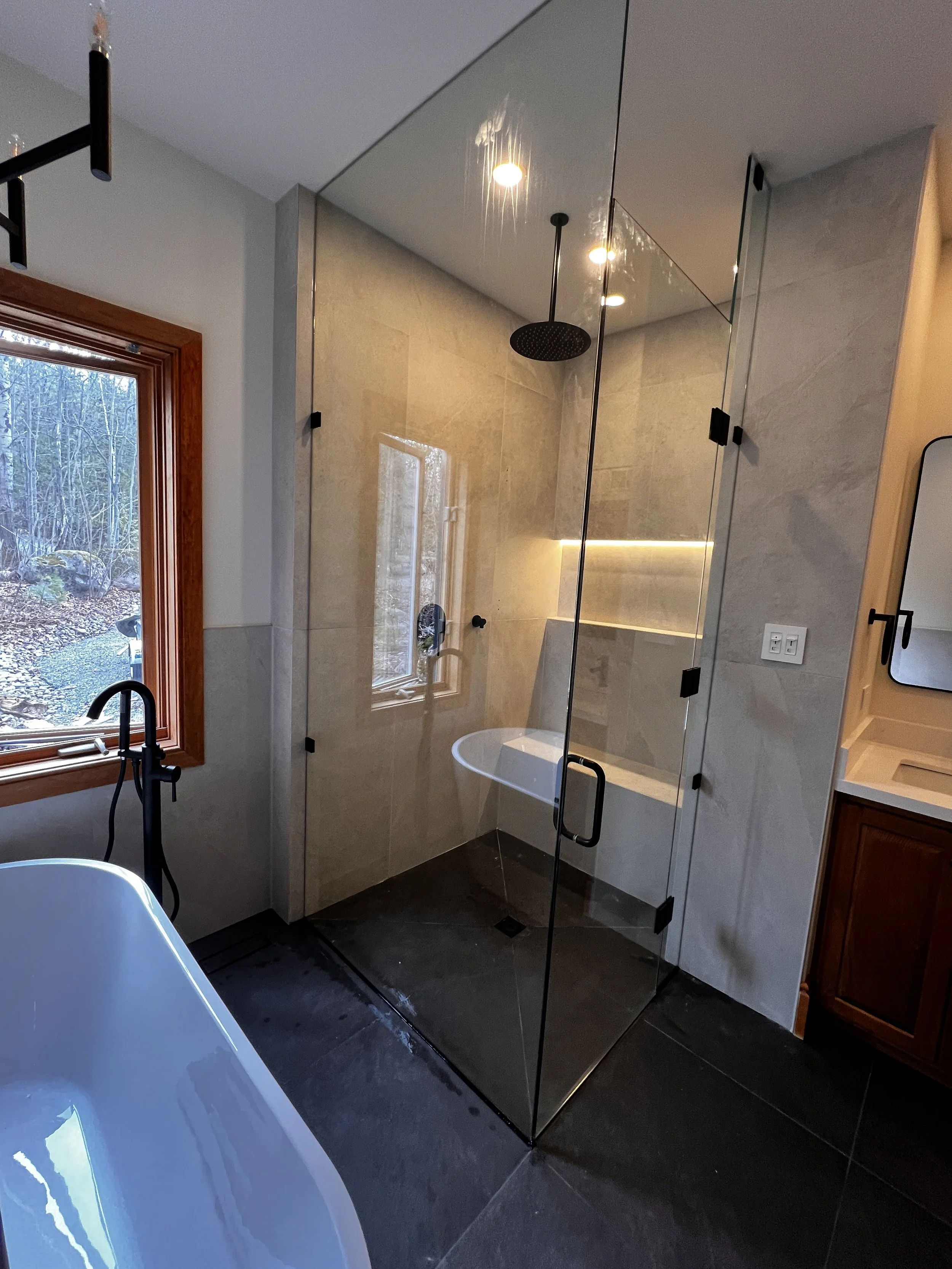 Modern bathroom with a glass shower enclosure, a bathtub next to a window, and a wooden vanity with a mirror.