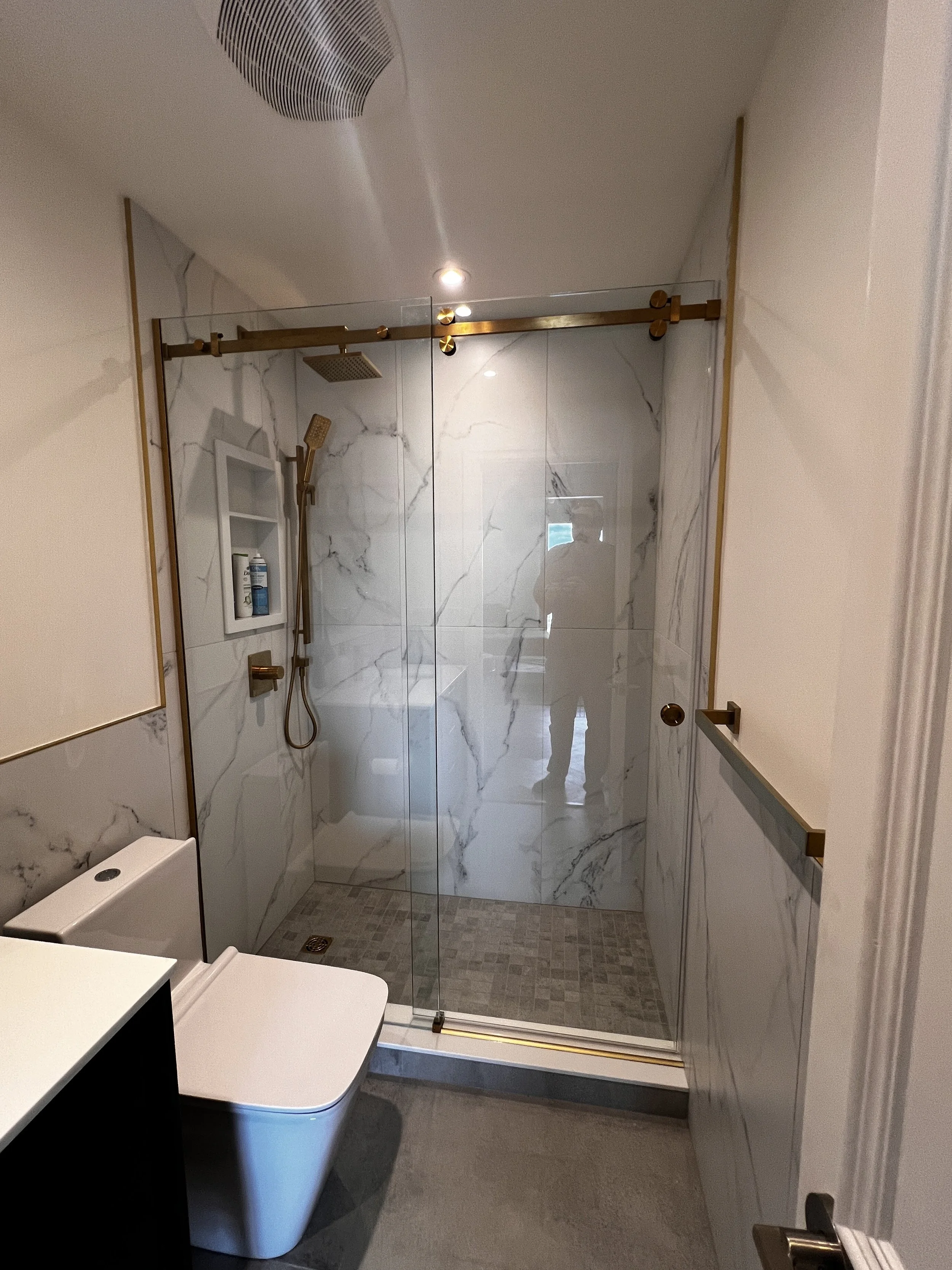 Modern bathroom with a glass-enclosed shower featuring marble walls, a gold sliding door track, a built-in shelf with toiletries, and a white toilet.