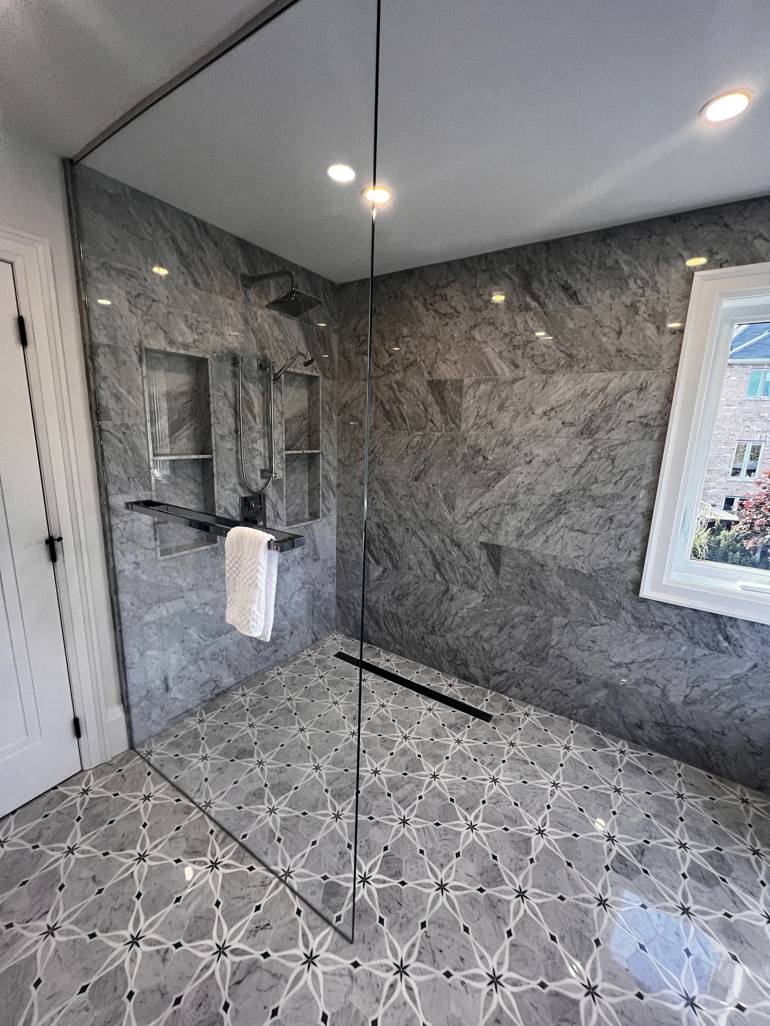 Modern bathroom with a glass shower enclosure, gray marble tiles on the wall, patterned tile floor, and a window with white trim.
