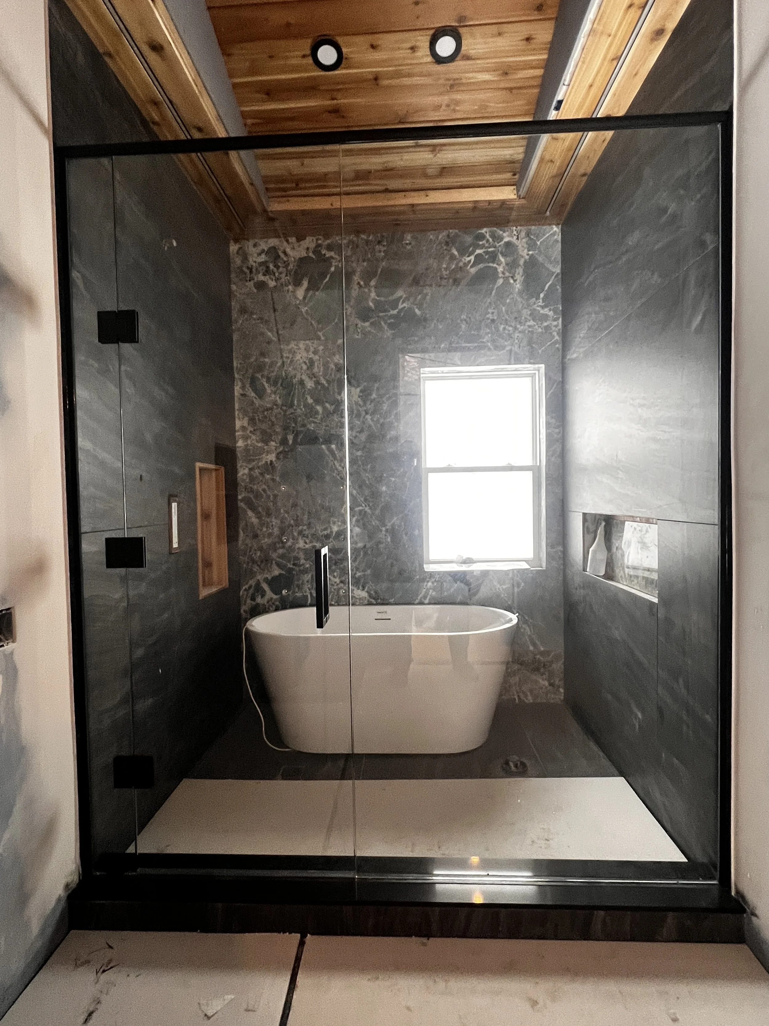 Modern bathroom with a walk-in shower, black framing, marble walls, a freestanding bathtub beneath a window, and a wood-paneled ceiling.