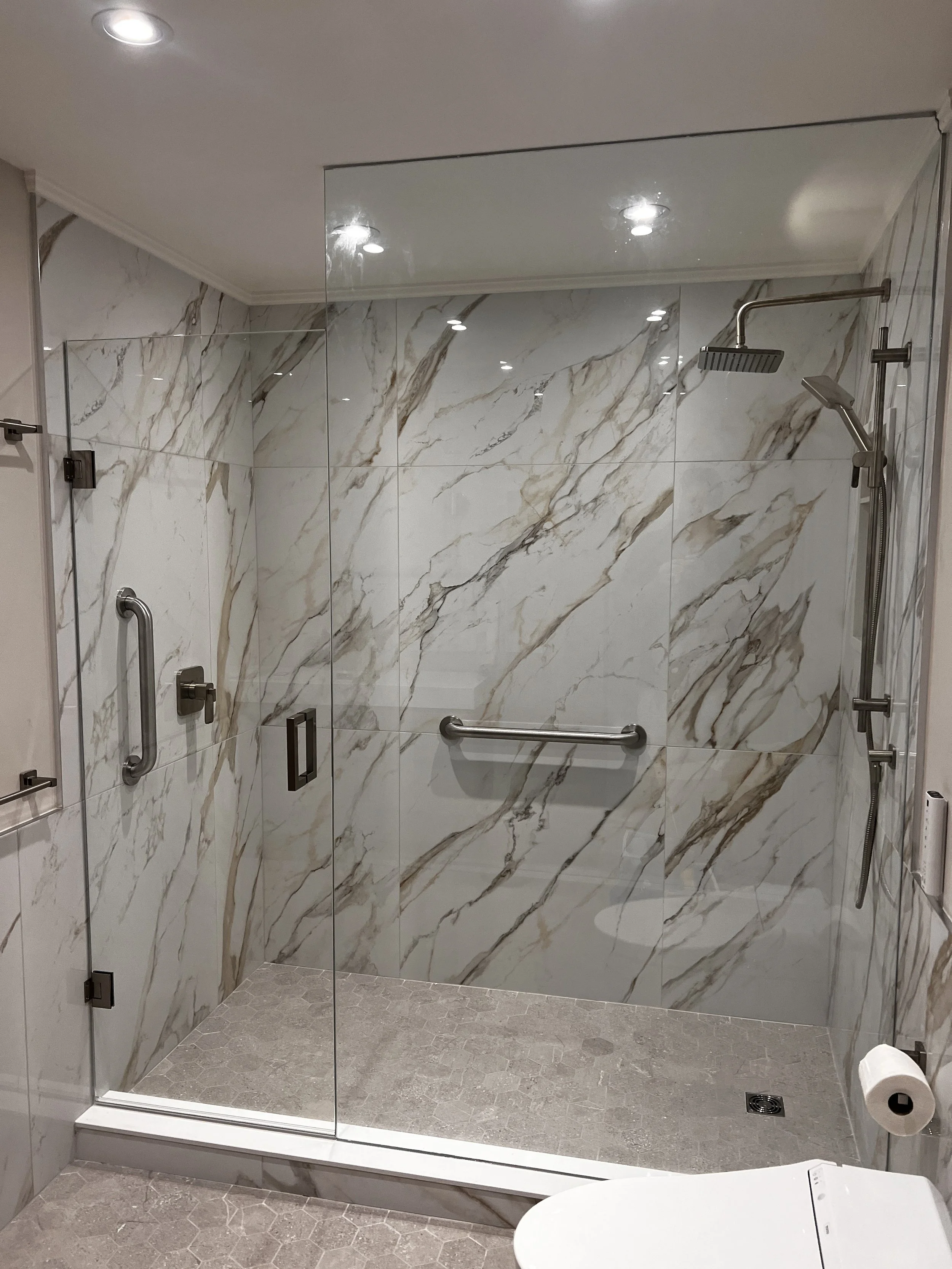 A modern walk-in shower with glass doors and marble tiles, featuring a rain showerhead, a handheld shower, and a grab bar. Toilet paper is visible in the bottom right corner.