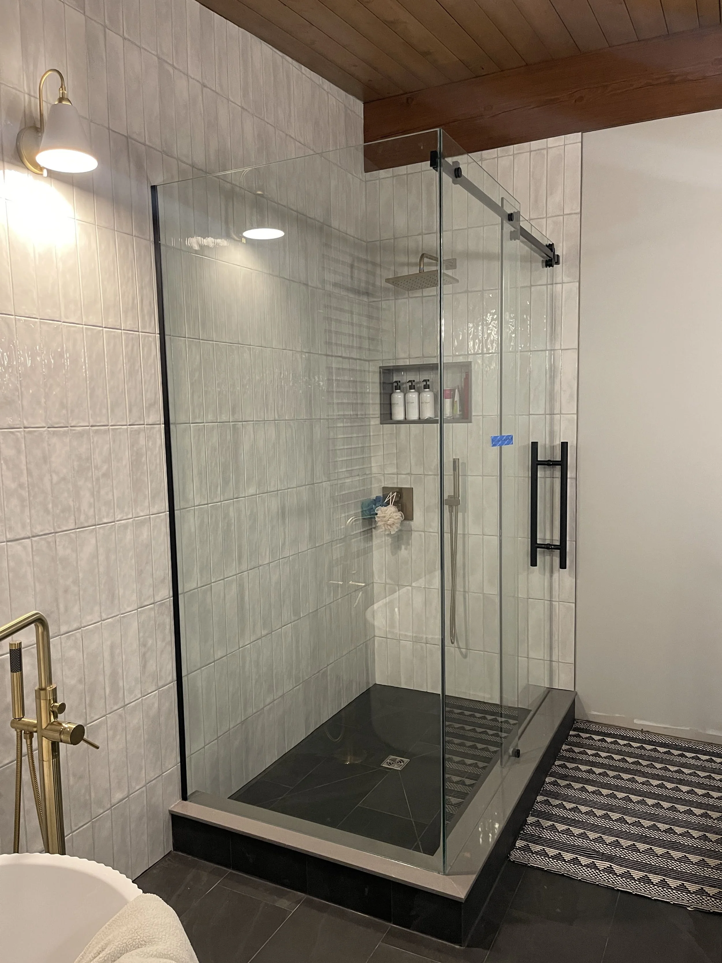 A modern walk-in shower with glass enclosure, black handle, rain showerhead, wall niche with toiletries, and black floor tiles.