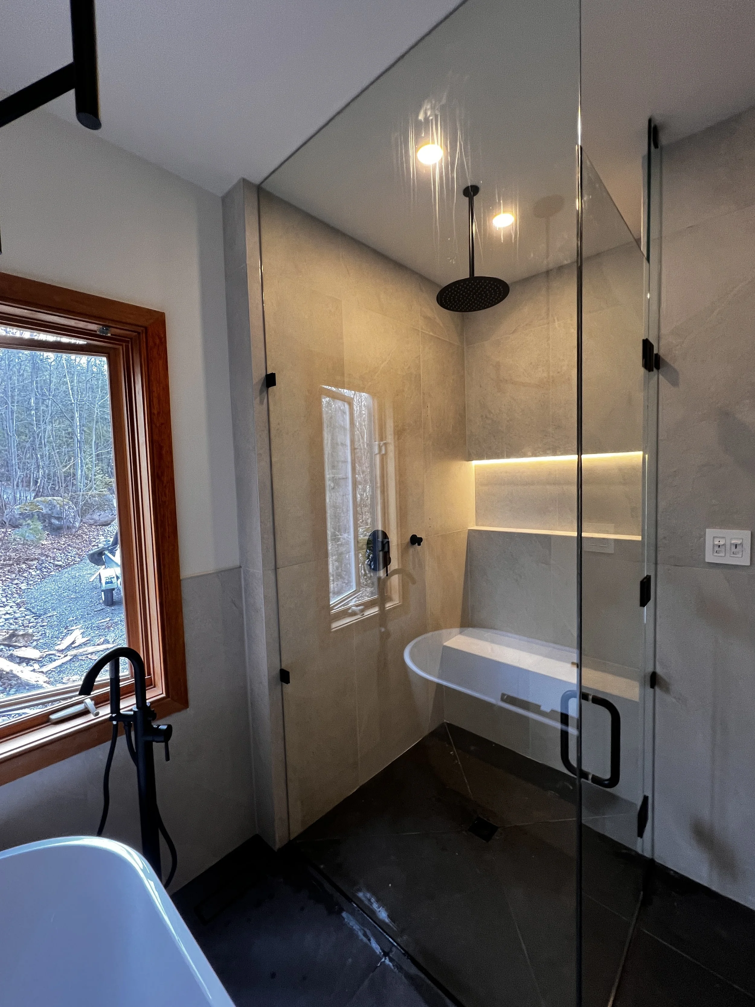 Modern bathroom with a walk-in shower featuring a black rainfall showerhead, a small window, and a bathtub inside the shower enclosure. Natural light from a large window with a wooden frame illuminates the space.