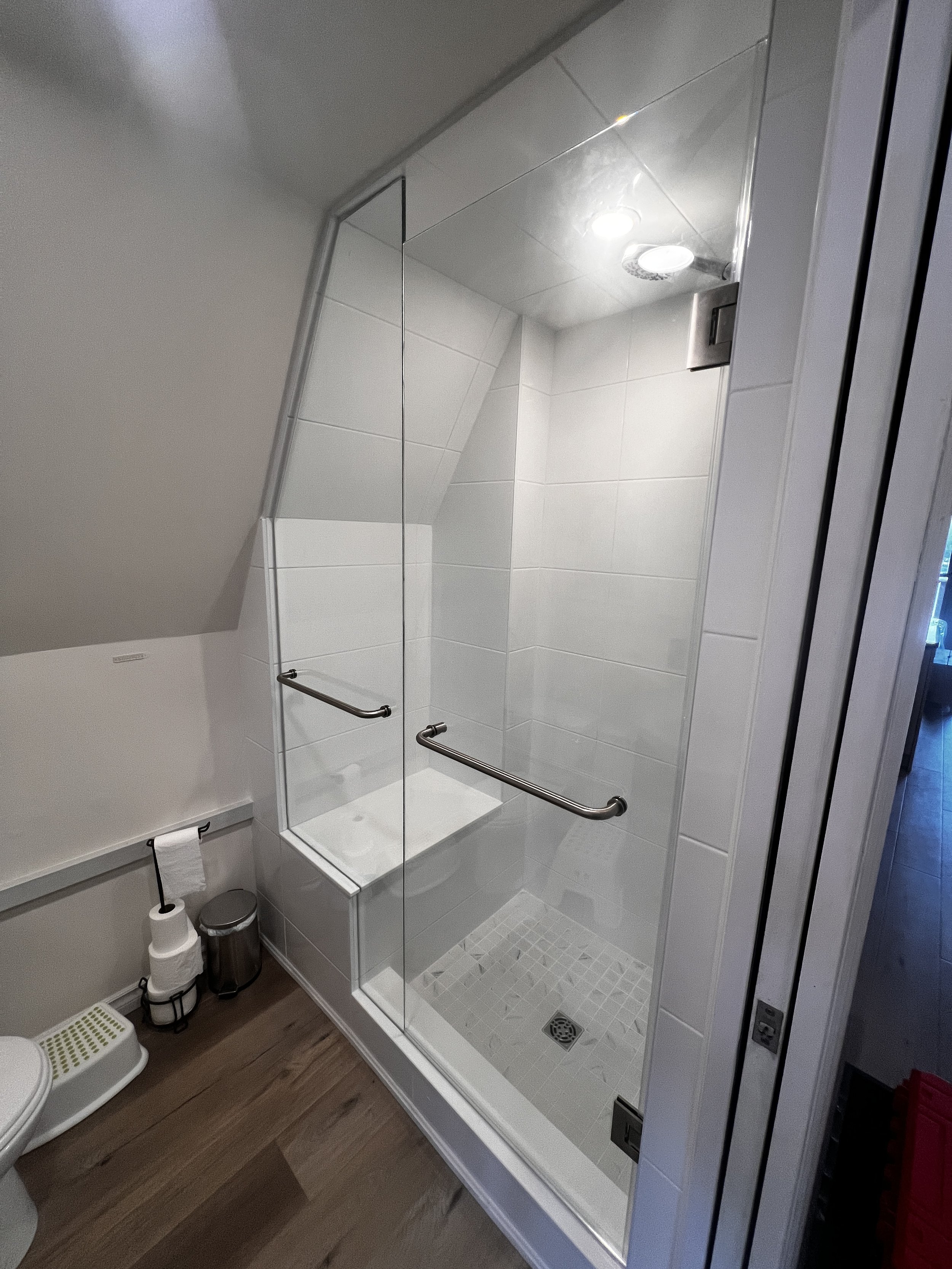 A walk-in shower with a glass door, white tiled walls, and a shower drain on the floor, with a grab bar inside. To the left, there is a toilet, toilet paper, a trash bin, and a step stool on a wooden floor.
