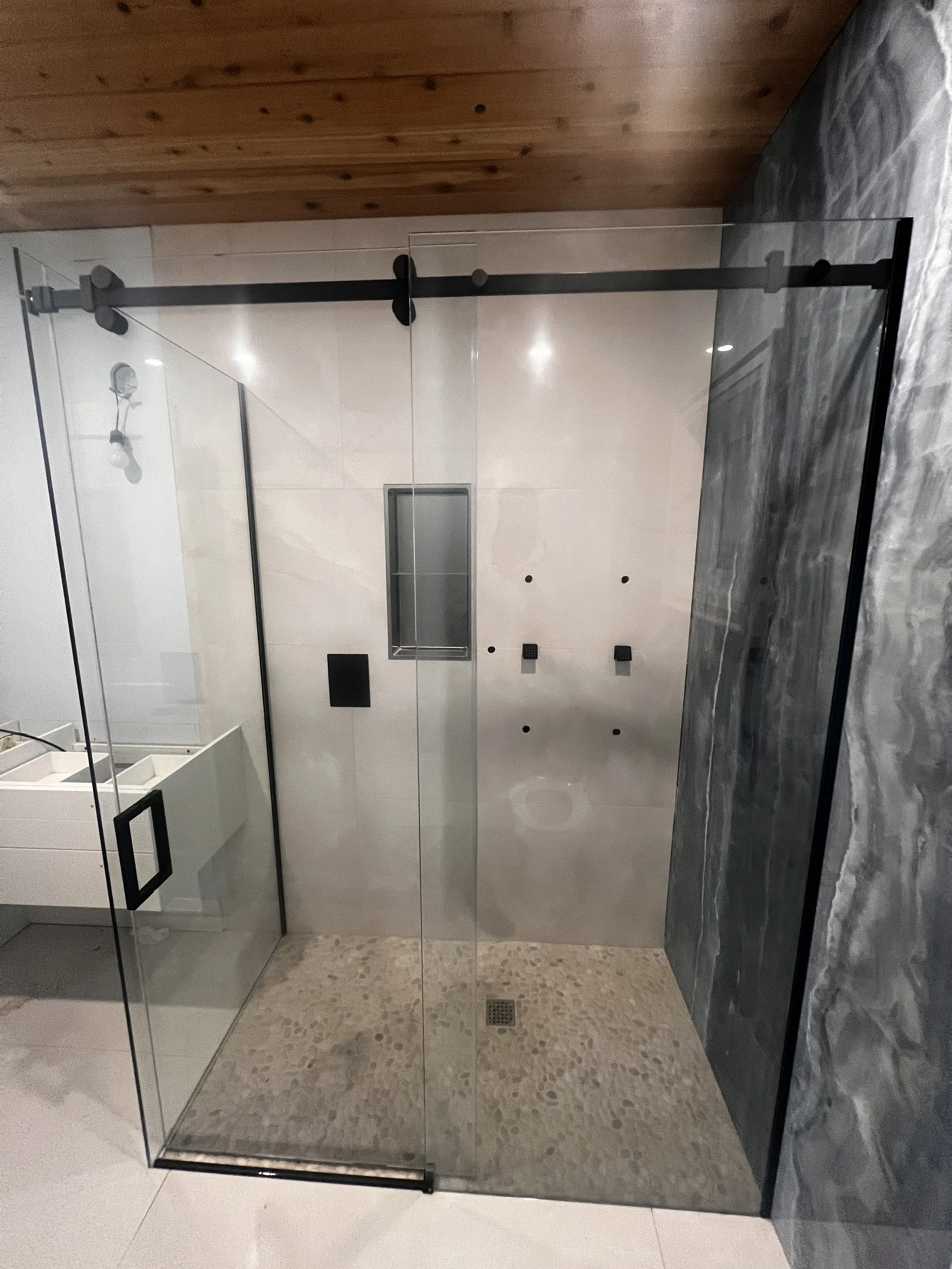 Modern shower with glass sliding doors, stone floor, gray marble wall, and ceiling wood paneling.