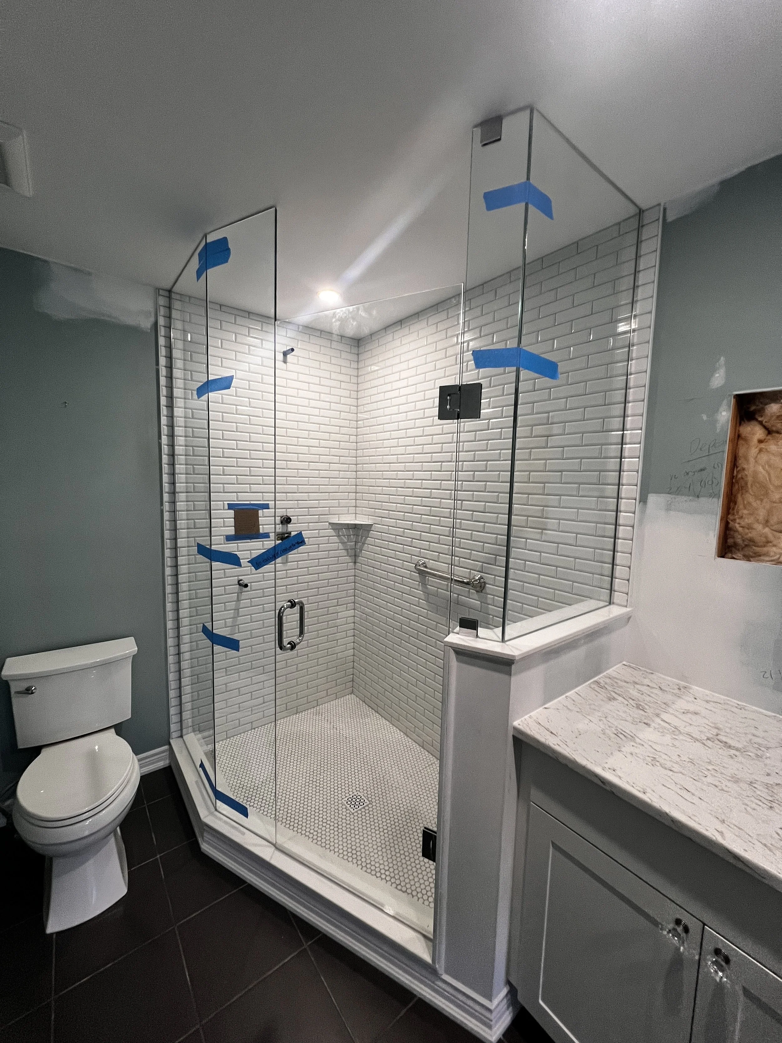 Newly installed glass shower enclosure with white subway tile walls, small hexagonal floor tiles, and a built-in corner shelf. Bathroom has a toilet on the left and a gray vanity with a granite countertop on the right.