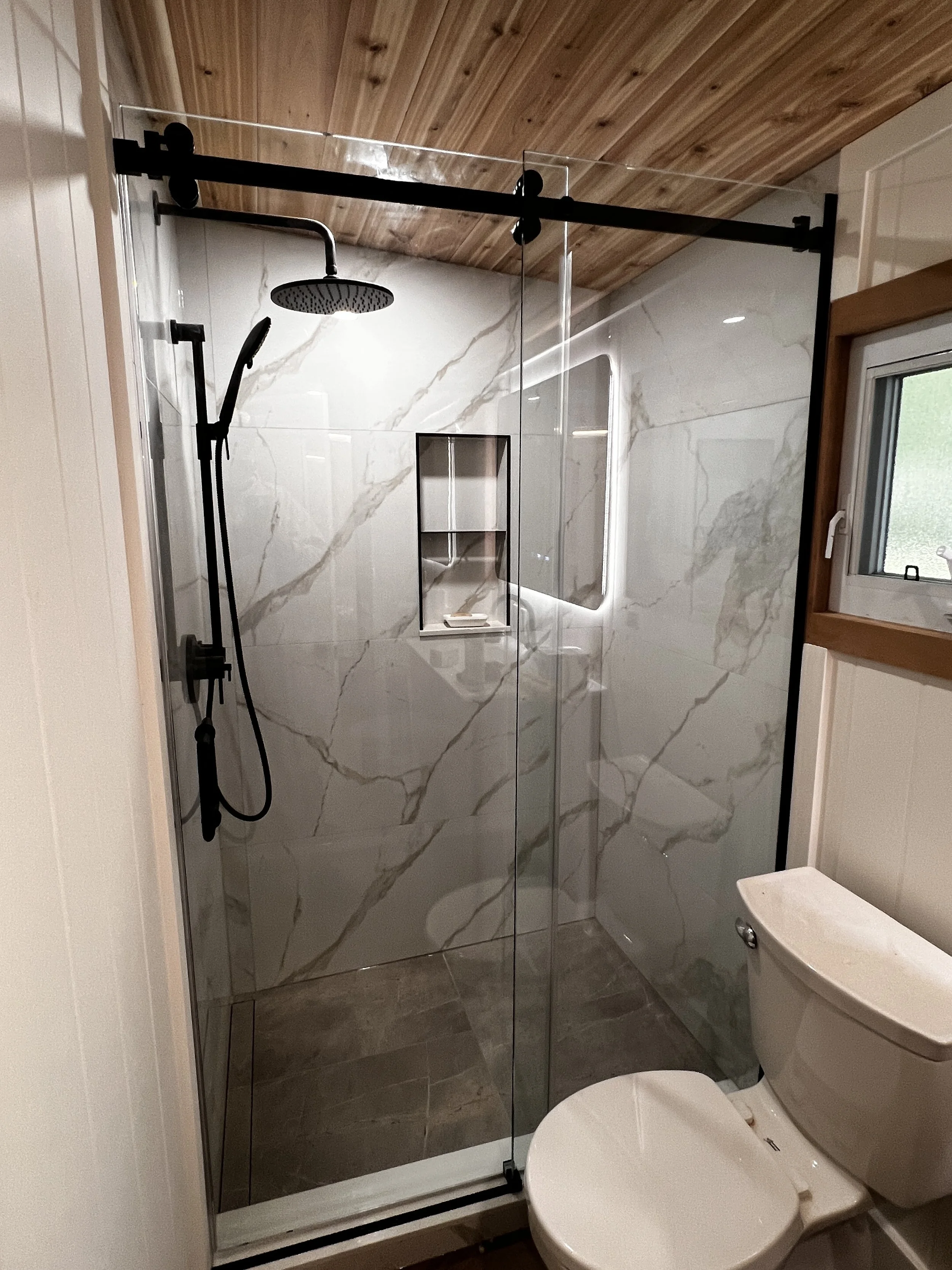 A modern bathroom with a marble-tiled shower with black fixtures, a built-in shelf, a rain shower head, and a hand-held shower wand, next to a white toilet and a small window with a wooden frame.