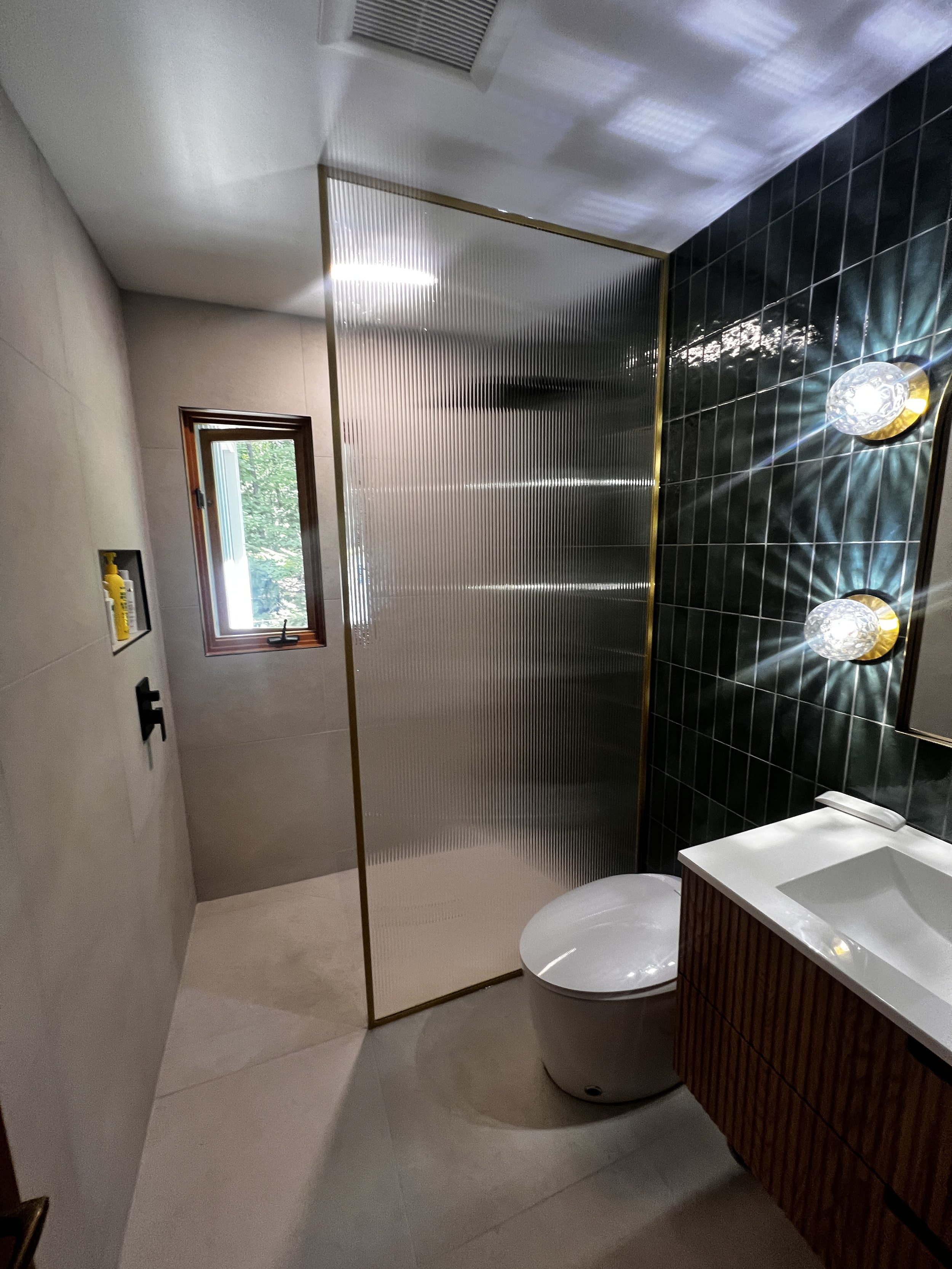 A bathroom with a small window, black tiled wall, and a glass shower enclosure with a gold frame. There is a modern white toilet, a wooden vanity with a white sink, and two decorative wall-mounted light fixtures.