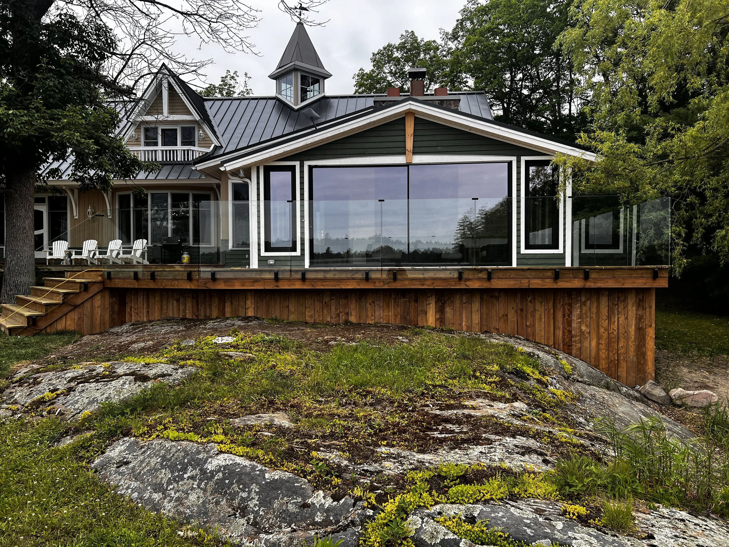 A house with a large glass enclosed porch or sunroom on a wooden deck, with chairs on the side, surrounded by greenery and rocks in the yard.