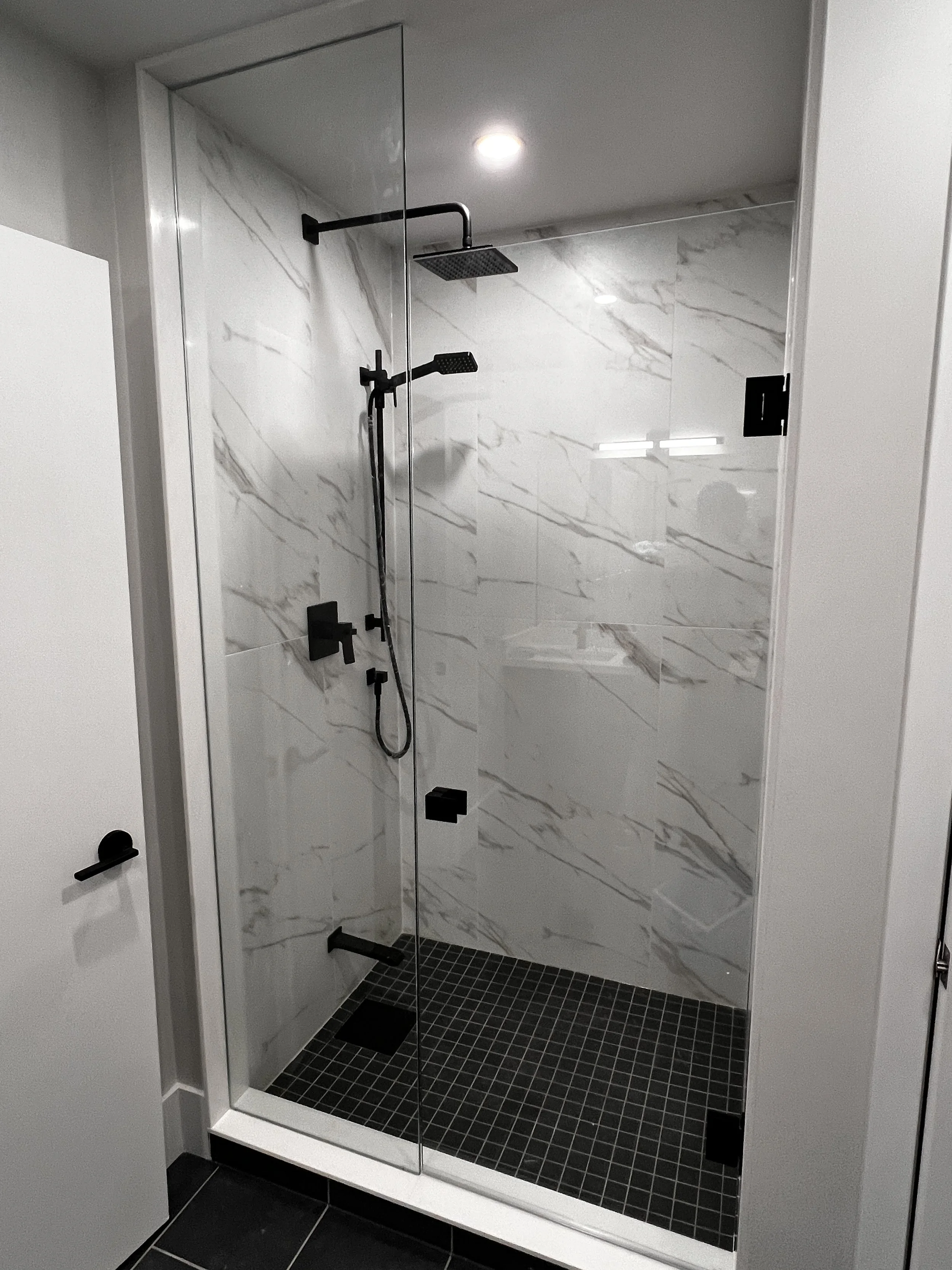 Modern shower with black fixtures, white marble tiled walls, black square floor tiles, glass enclosure, and overhead lighting.