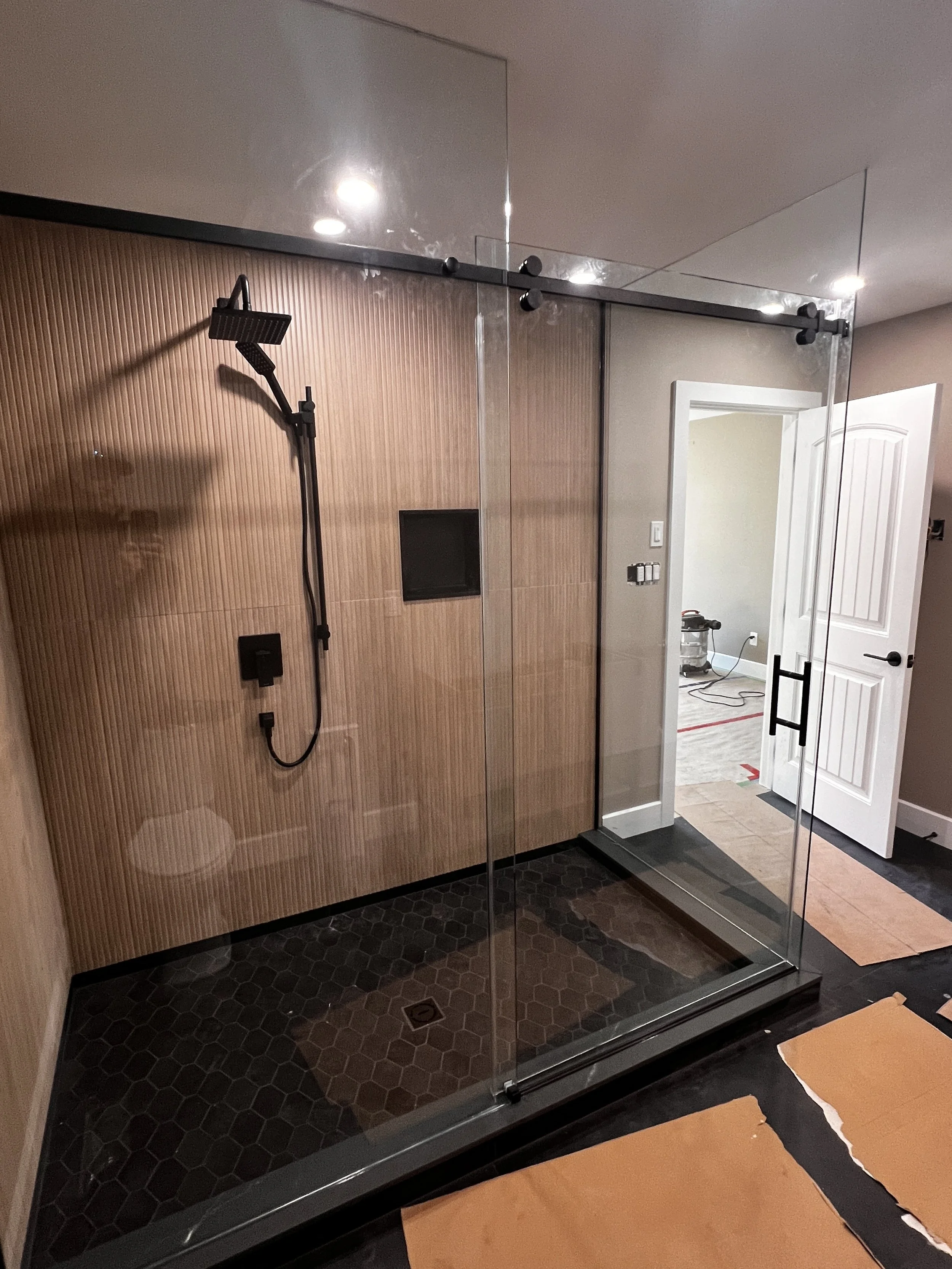A modern bathroom with a glass shower enclosure, black hexagon tile floor, wooden paneled wall inside the shower, black showerhead, and a small built-in shelf. A white door opens into an adjacent room with a vacuum cleaner and construction tools visible.