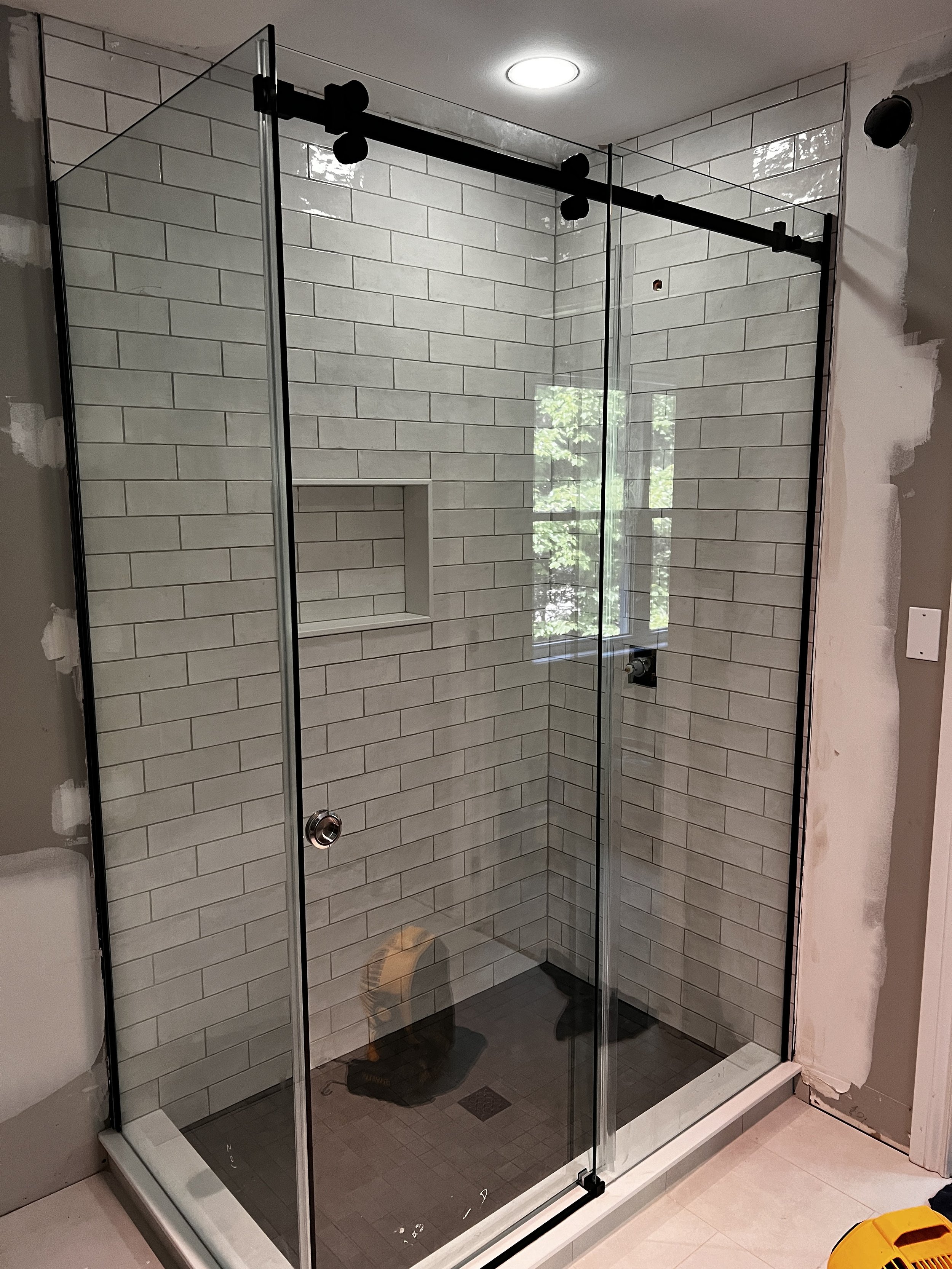 Glass shower enclosure with black sliding barn door hardware, tiled walls with a built-in niche, and a window reflecting outside greenery.