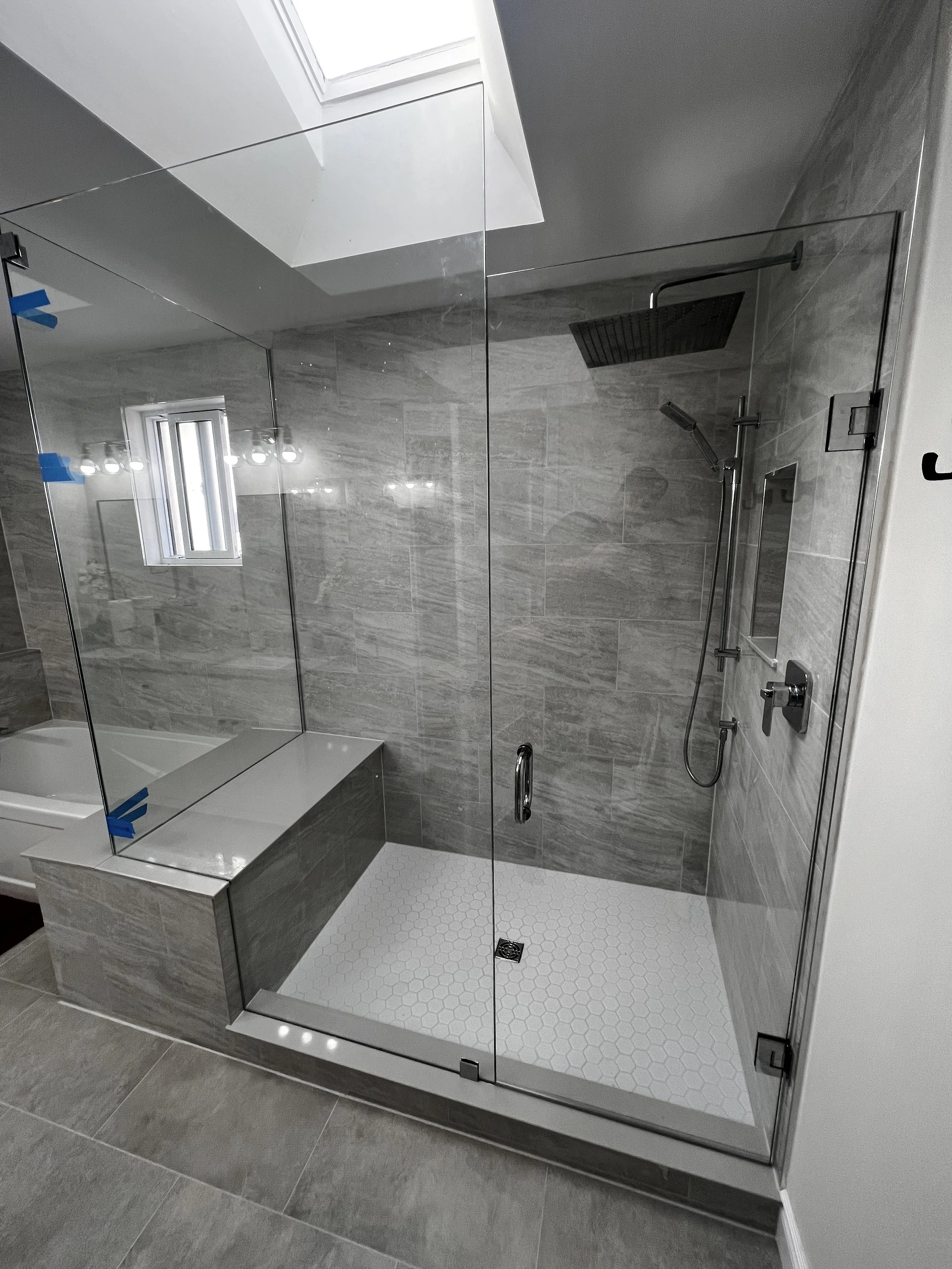 Modern walk-in shower with glass doors, gray tiled walls, white hexagonal floor tiles, a black square rain showerhead, a handheld shower, and a small built-in shelf.