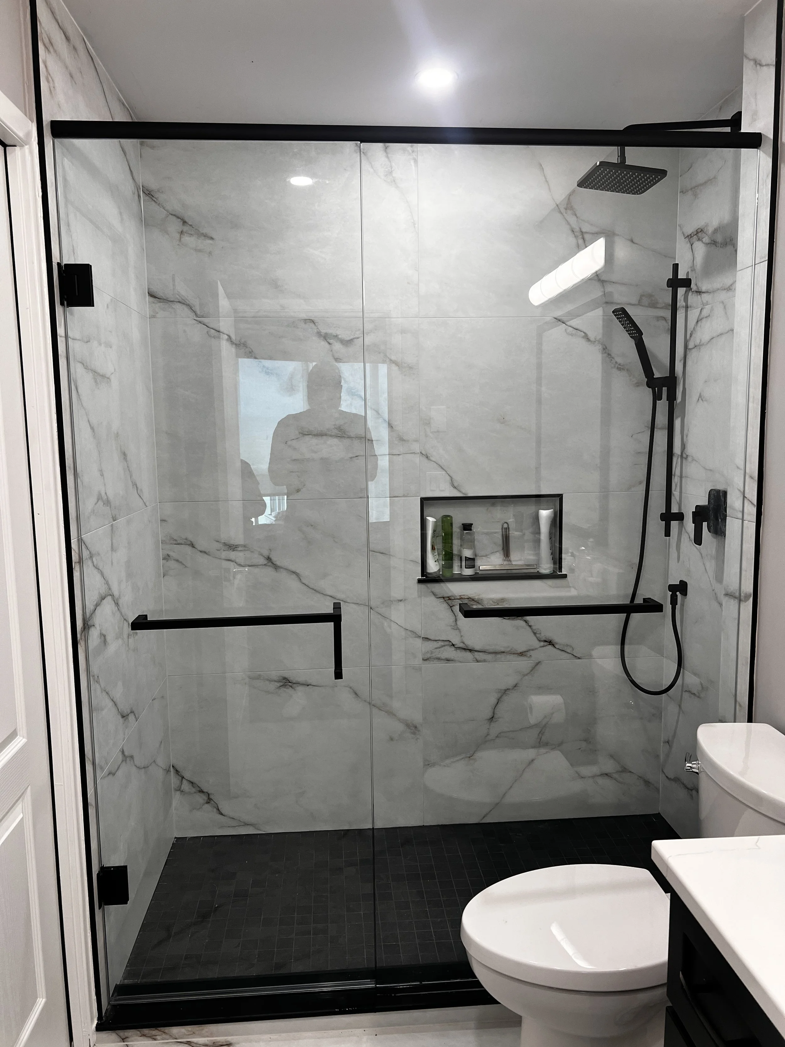 A modern bathroom with a frameless glass shower enclosure, white marble walls with gray veining, black fixtures, and a toilet.