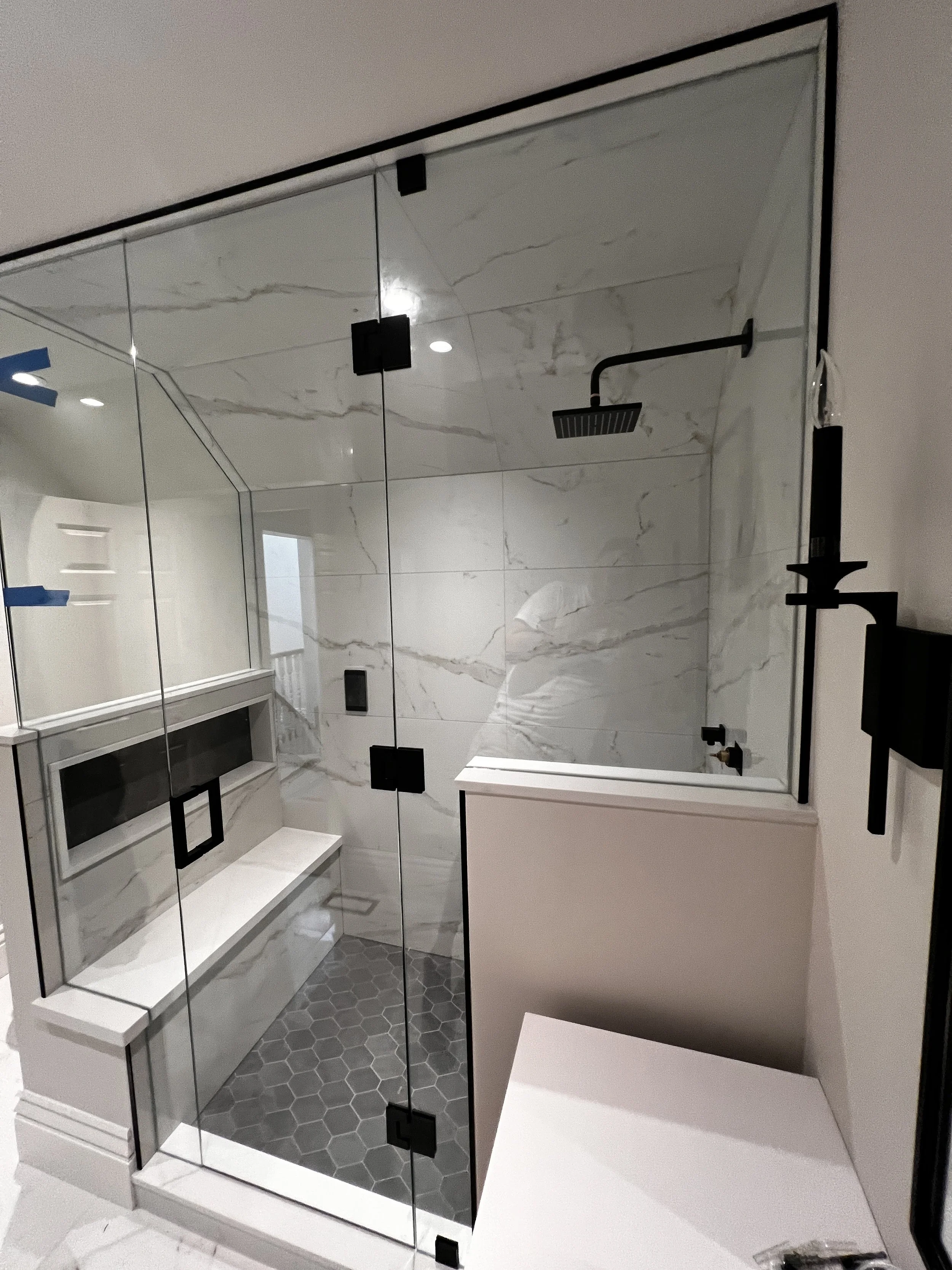 Modern bathroom with a glass shower enclosure, marble wall tiles, hexagonal floor tiles, a built-in bench, and black fixtures including a showerhead and candle holder.