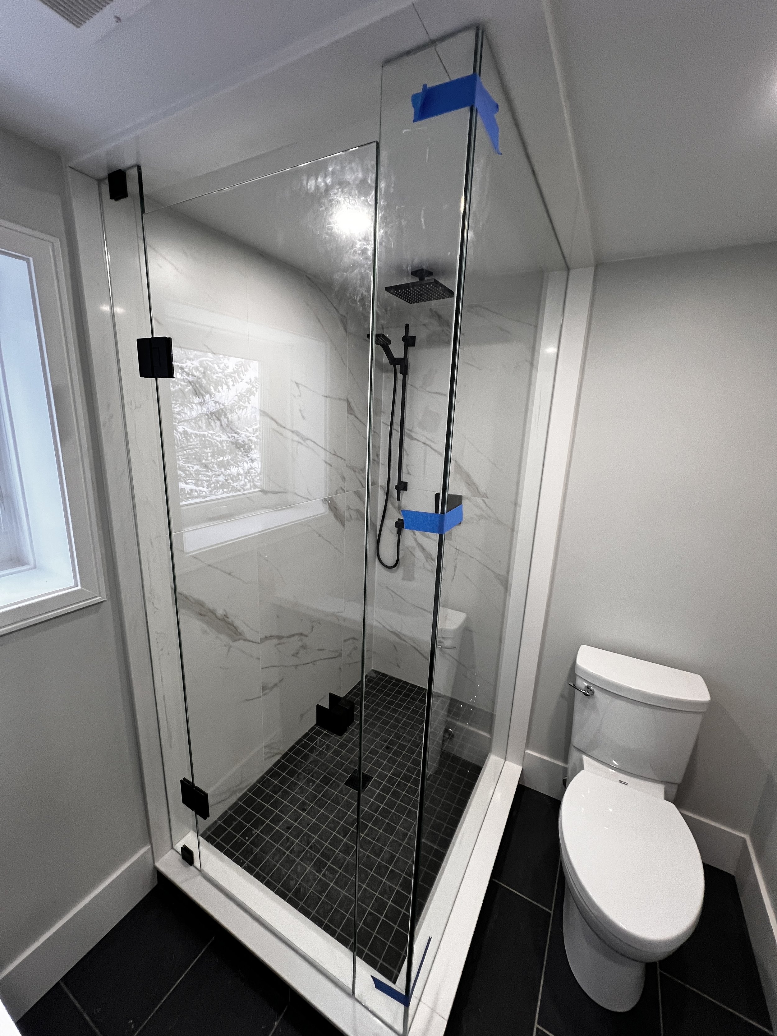 Modern shower with clear glass enclosure, black shower fixtures, and black tiled floor next to a white toilet in a bathroom with white walls and black floor tiles.