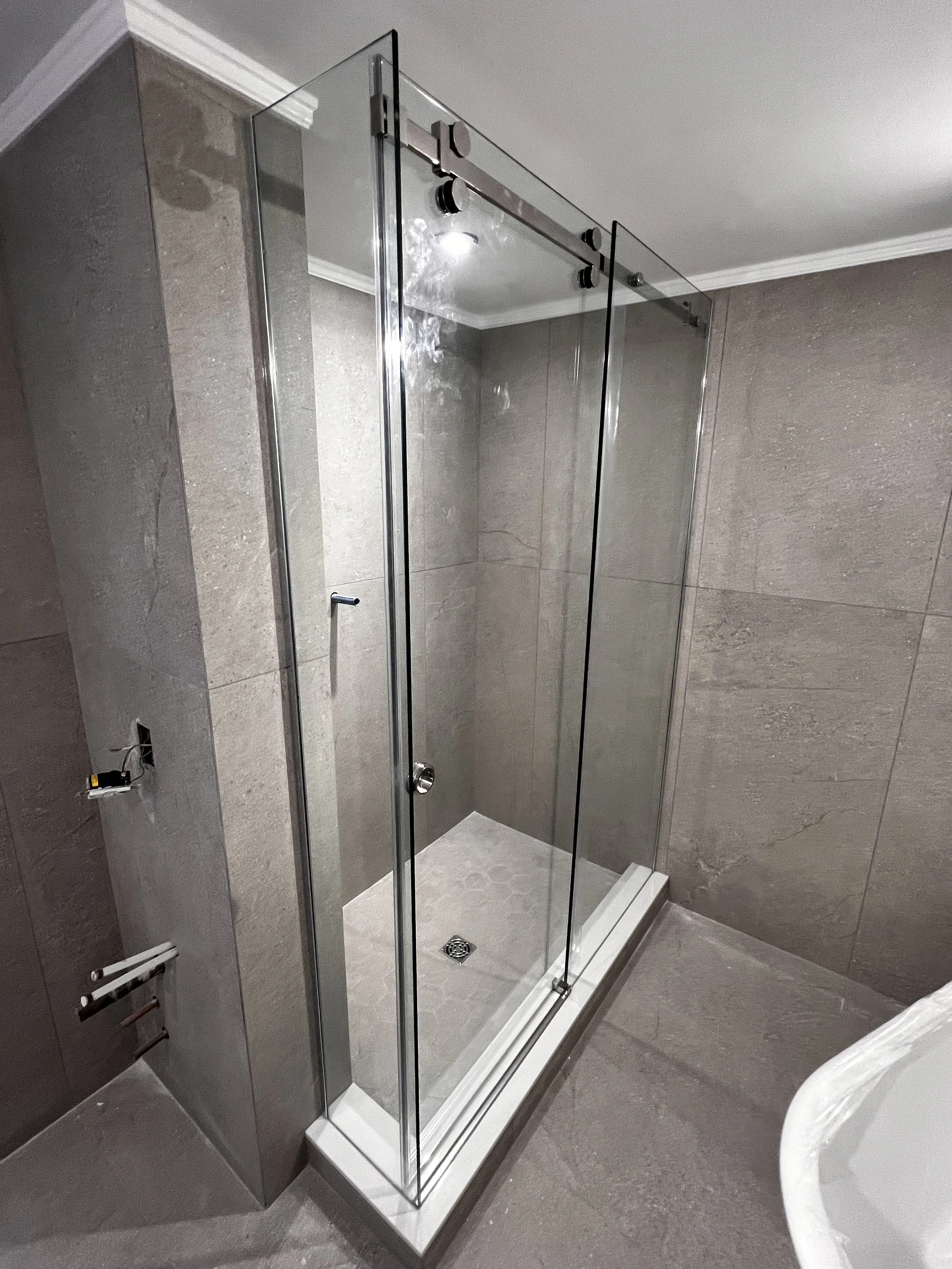 Empty modern glass shower enclosure with a chrome sliding door track in a bathroom with gray tile walls and floor, and a white bathtub partially visible in the foreground.