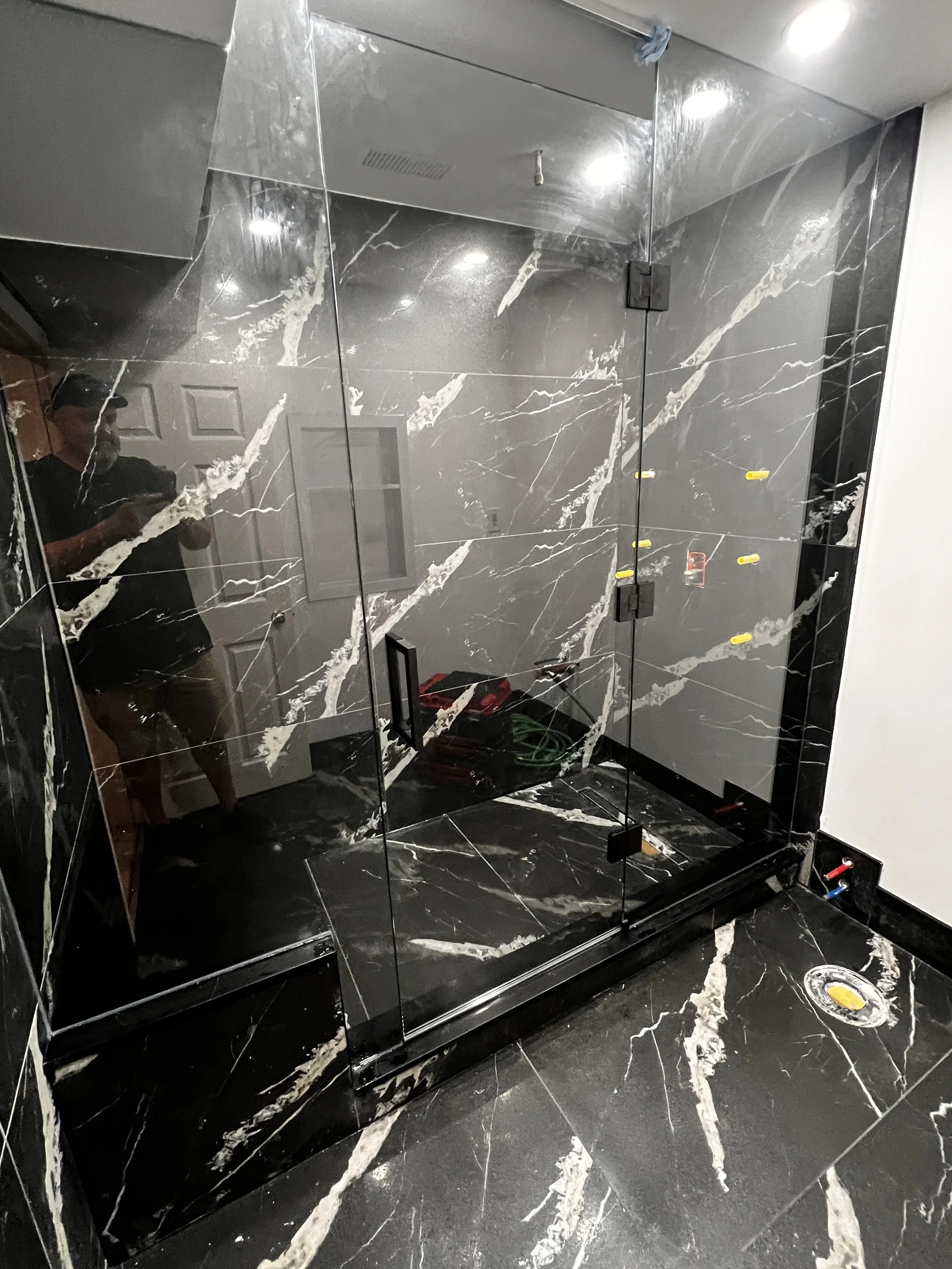 A black marble shower with glass doors, inside a bathroom under construction, with tools and piping visible.