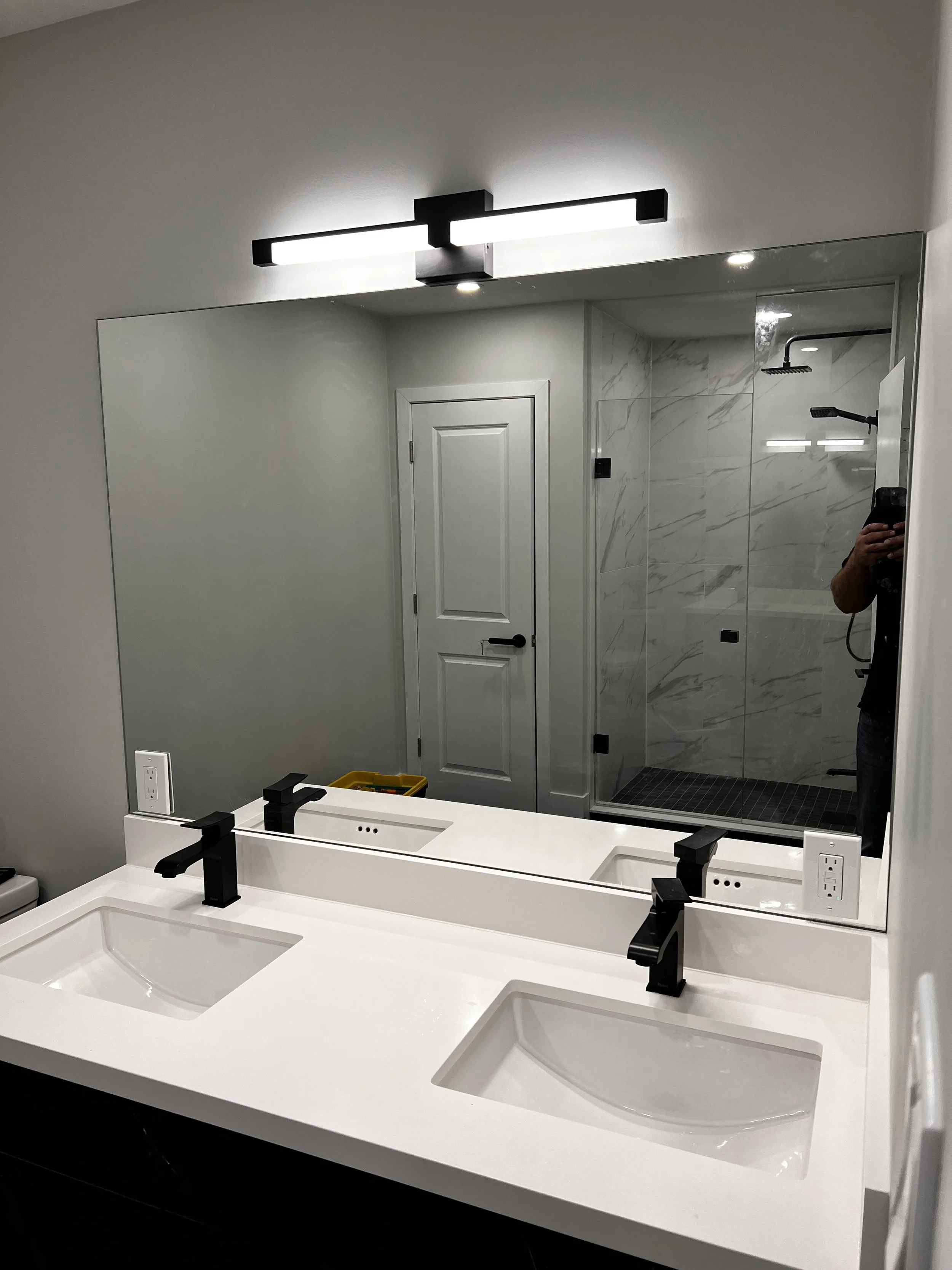 Modern bathroom with double vanity, black faucets, large mirror, and walk-in shower with glass doors and marble walls.