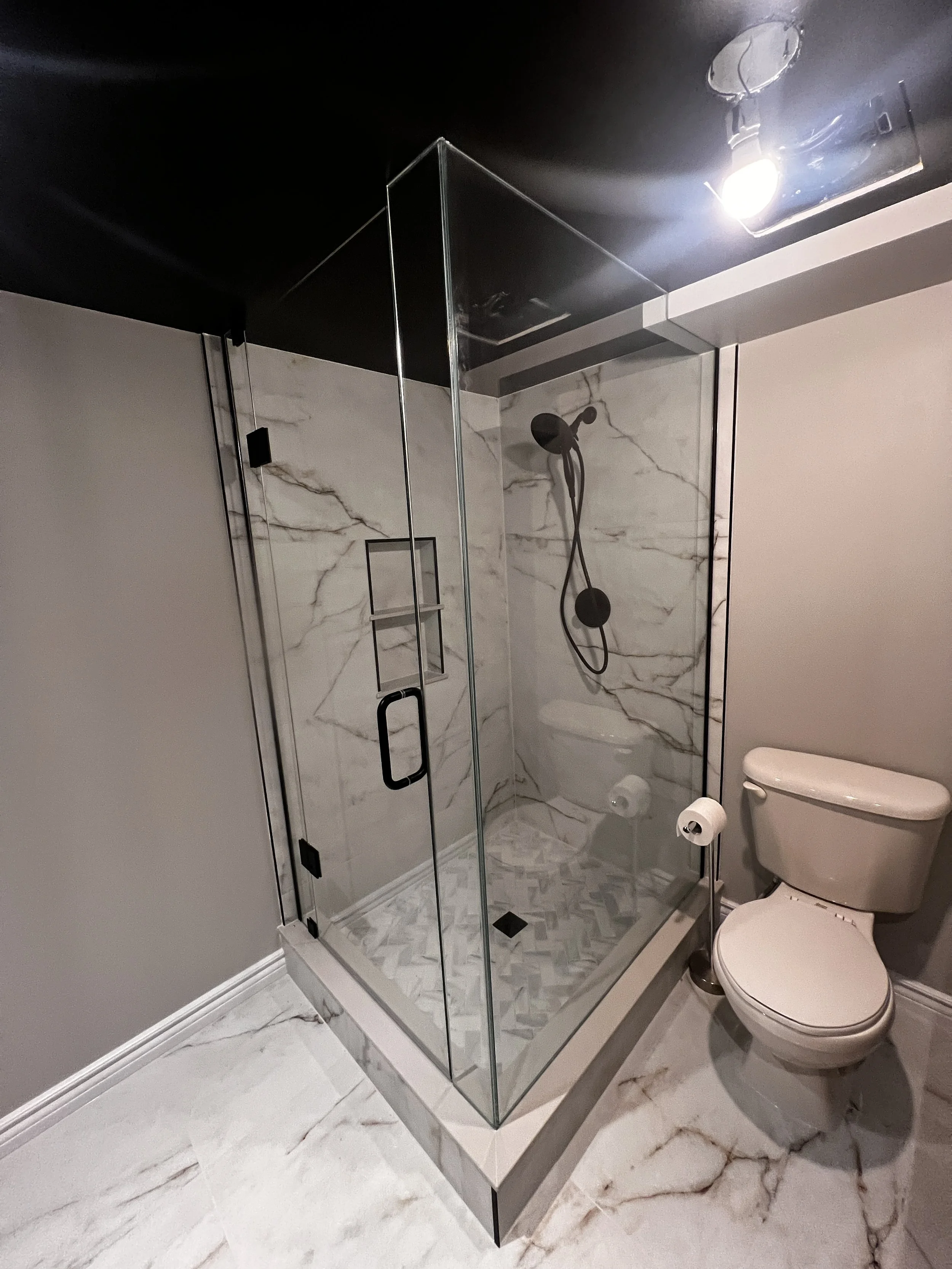 A modern bathroom with a glass shower enclosure featuring marble tile walls and flooring, a dark showerhead, and a white toilet beside the shower.