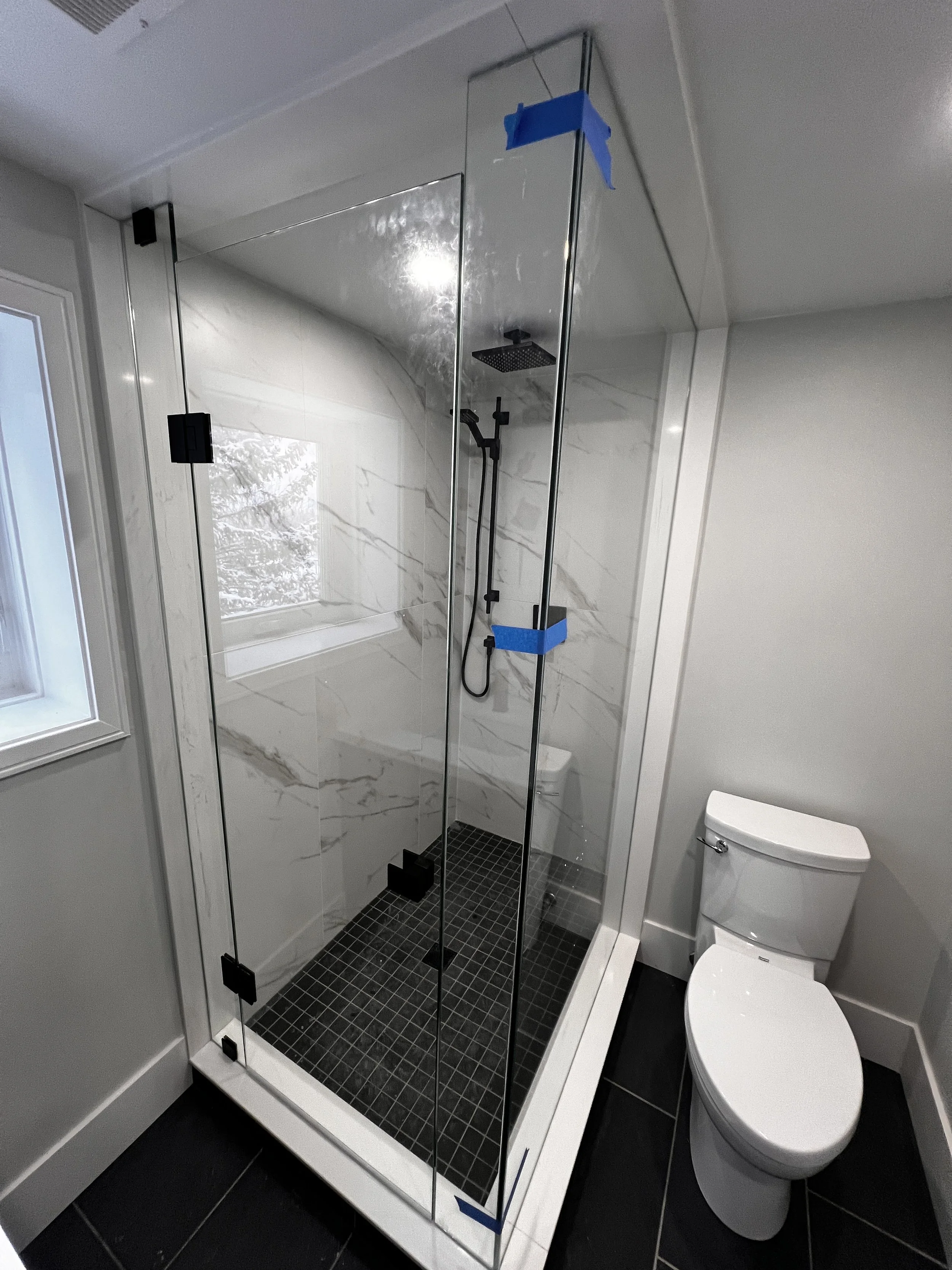 A modern bathroom with a glass shower enclosure and a white toilet, featuring black tile flooring and a marble wall inside the shower.