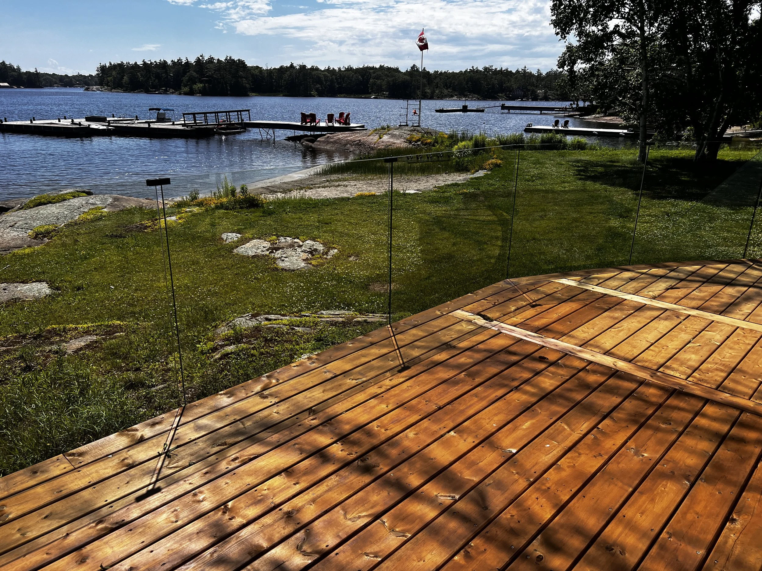 View of a lakeside area with a wooden deck in the foreground, a grassy shoreline, a dock with chairs and a Canadian flag in the middle ground, and trees and houses across the lake under a partly cloudy blue sky.