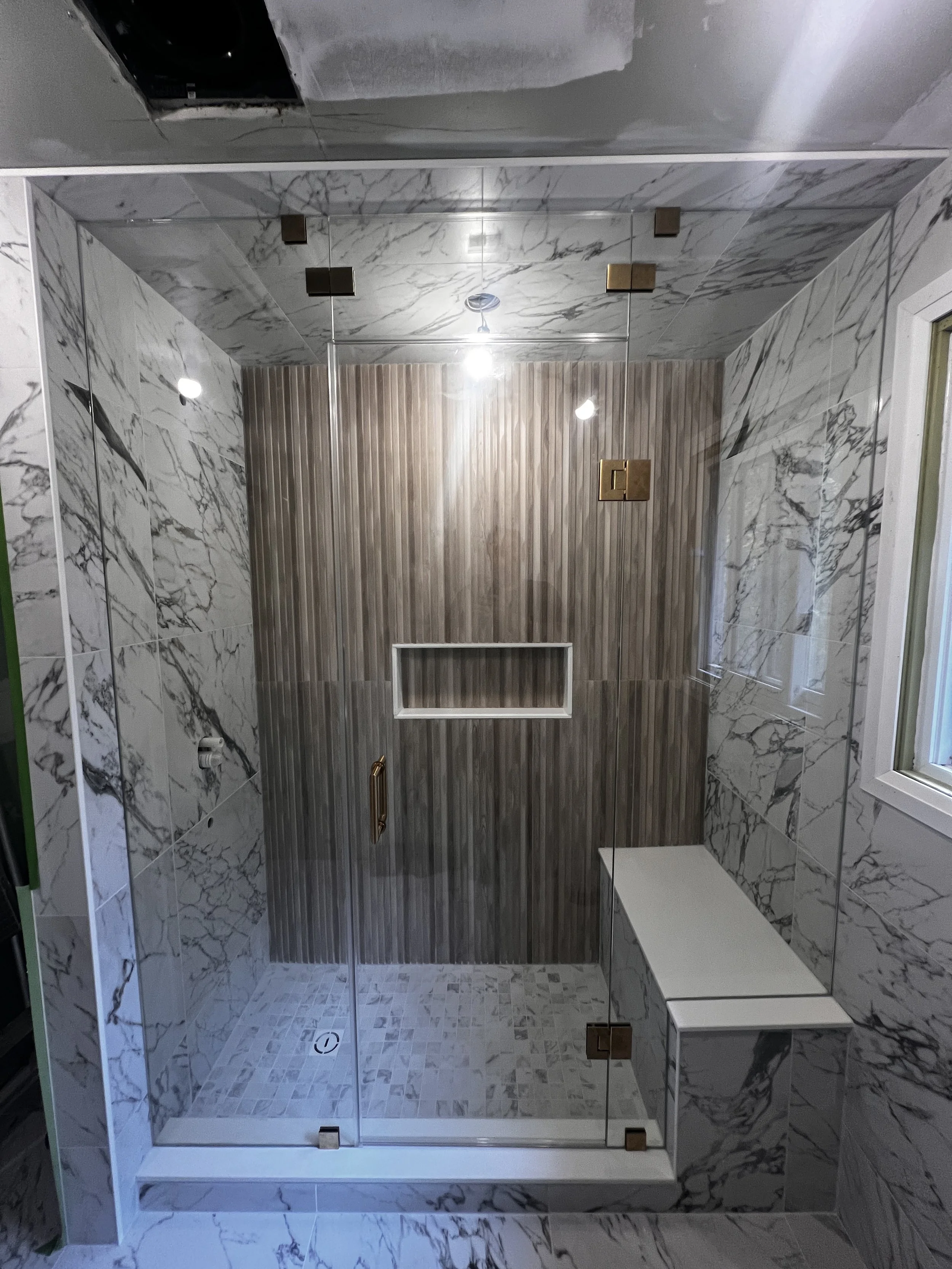 Modern walk-in shower with marble walls and floor, a wooden accent wall, glass doors with metal hardware, and a white bench.